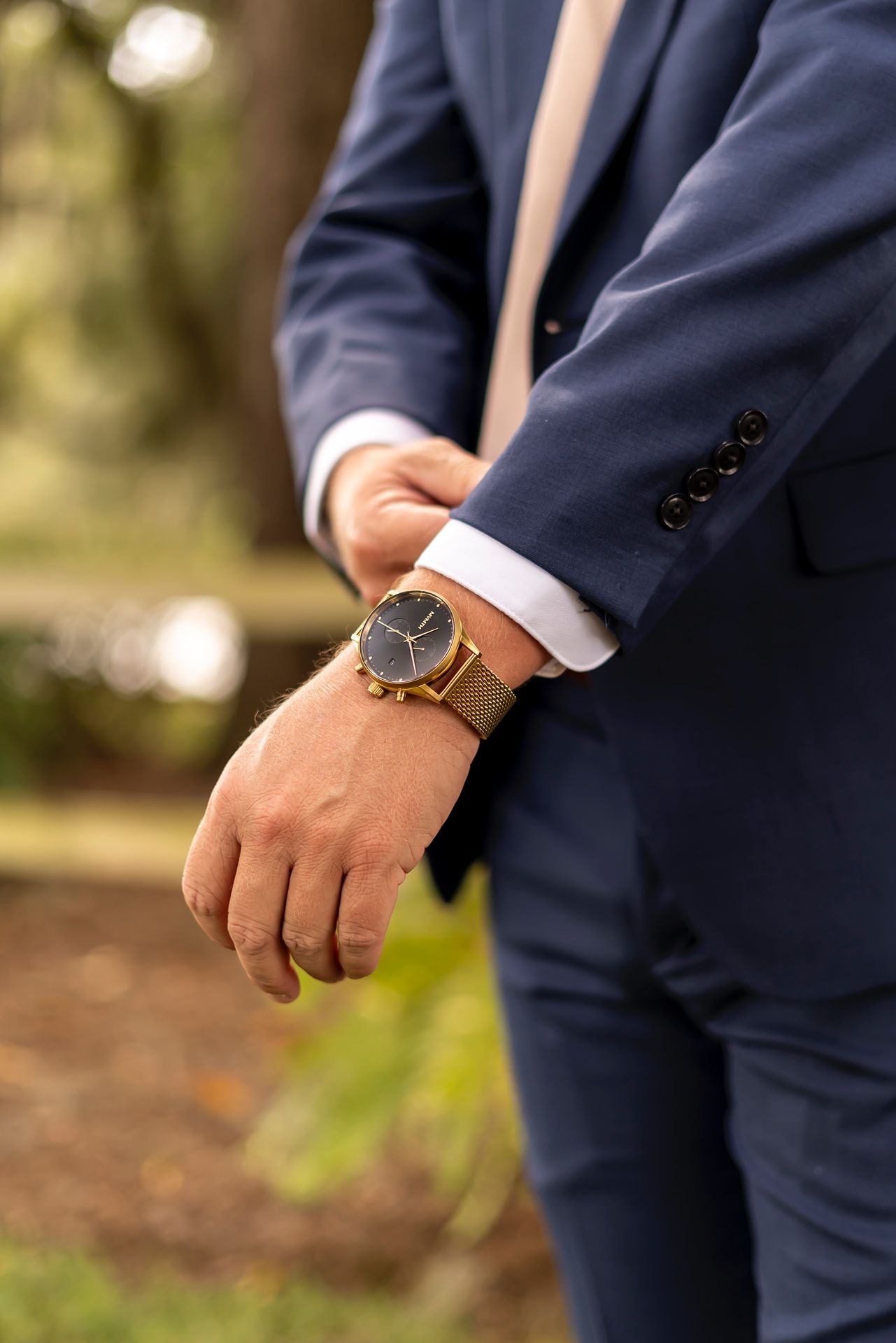 Close-up view of person wearing navy suit with brown leather watch on wrist against blurred outdoor background.