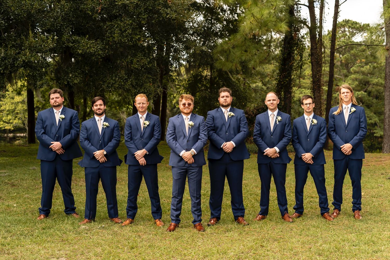 A wedding party in navy blue suits sit on a grassy hill during an outdoor photo session.