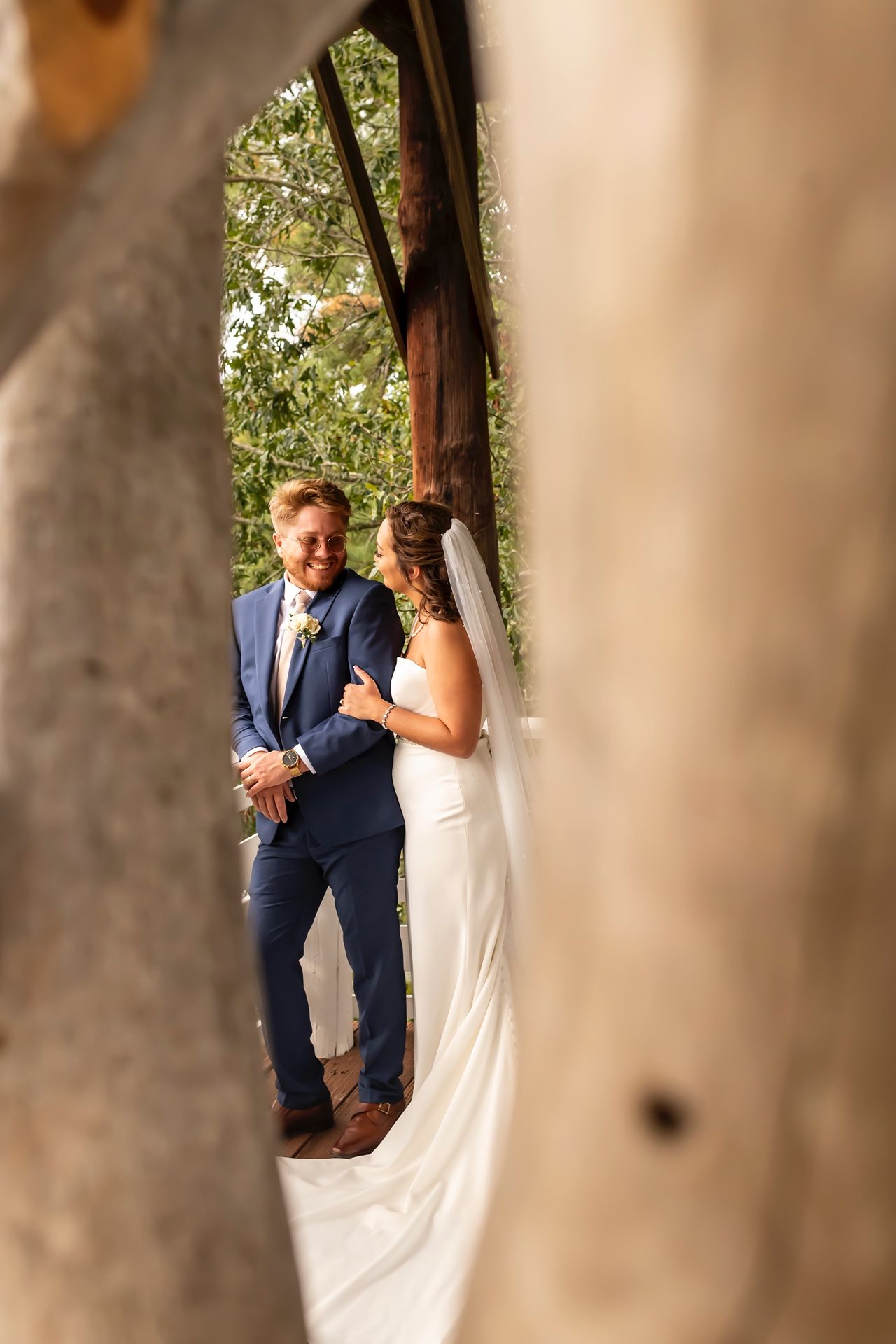 Couple in wedding attire shares intimate moment between rustic wooden pillars in outdoor setting.