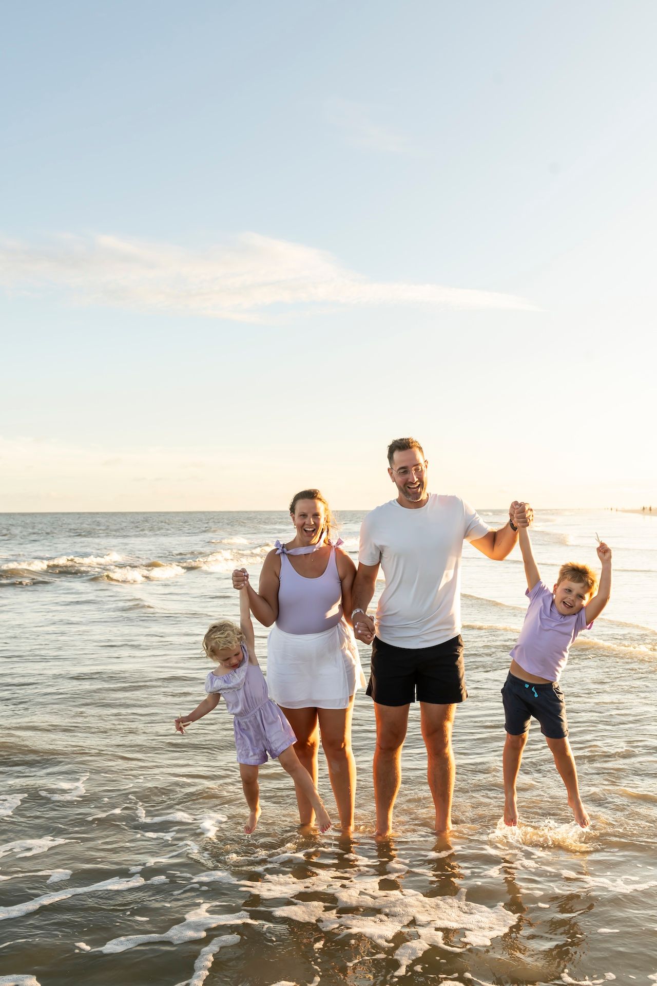 A family plays together in the ocean waves at sunset, wearing white shirts and dark shorts.