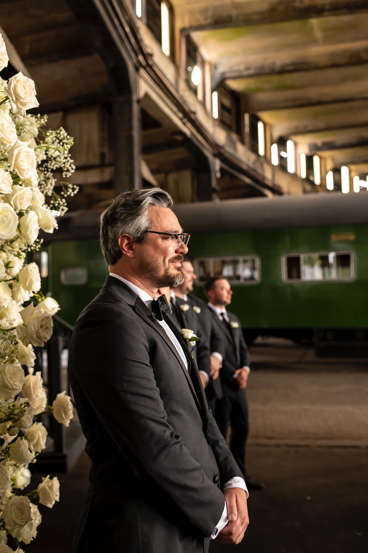 A formal wedding setup in an industrial venue with white floral arrangements and guests in black tuxedos.