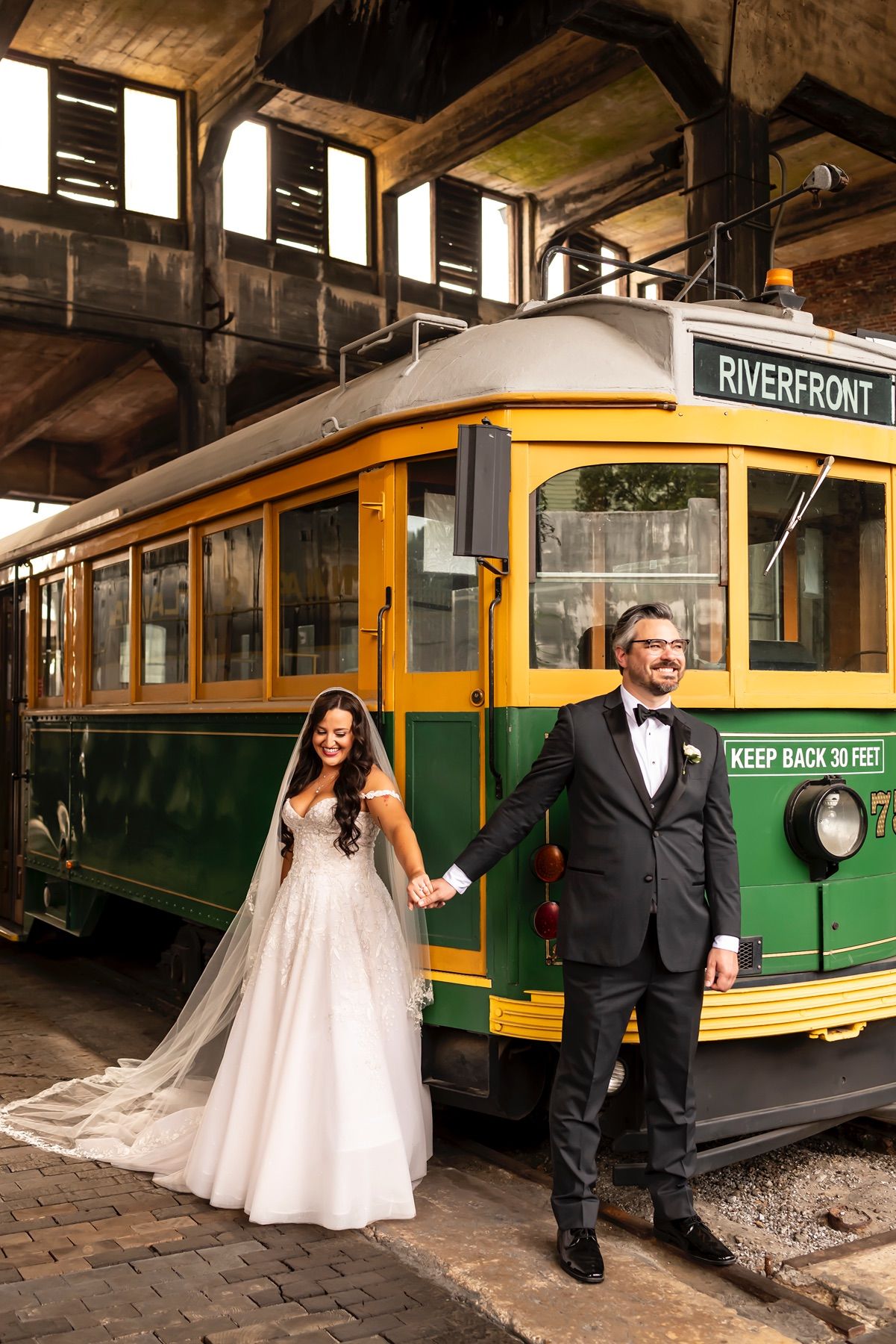 Vintage green and yellow Riverfront streetcar with couple in wedding attire standing beside it under covered station.