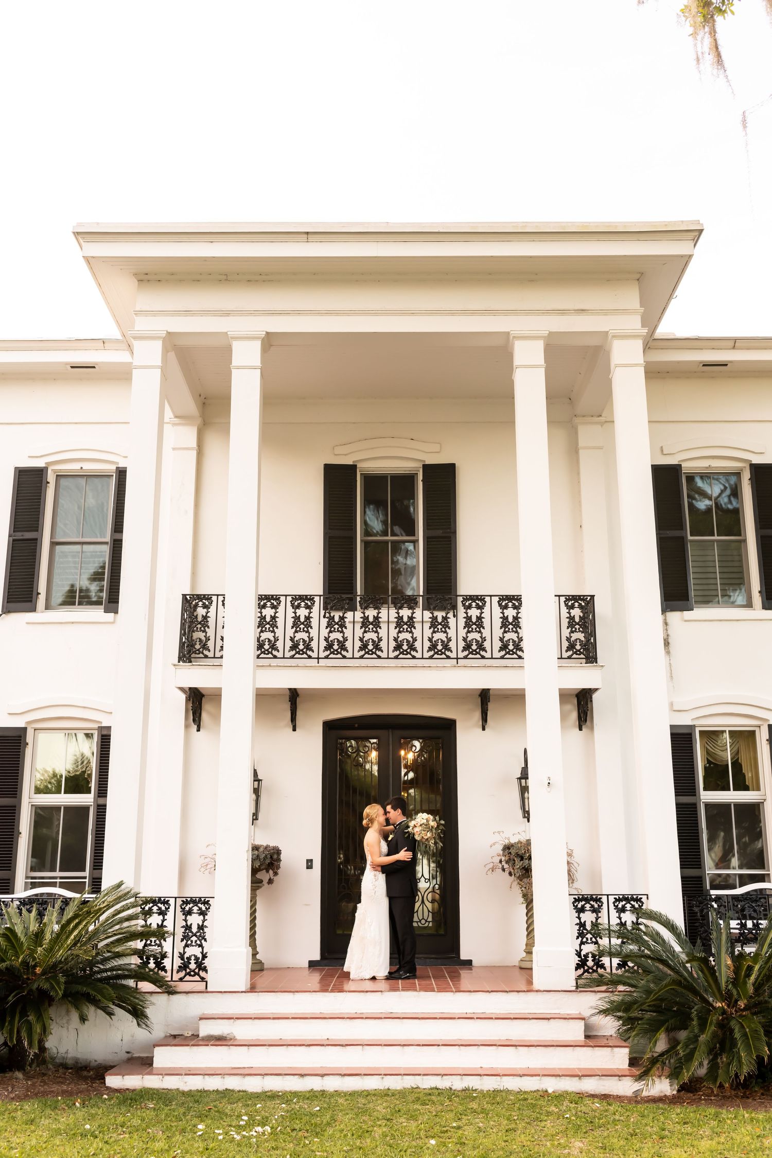 Bride and groom white antebellum mansion with grand columns and ornate iron railings on the front porch.