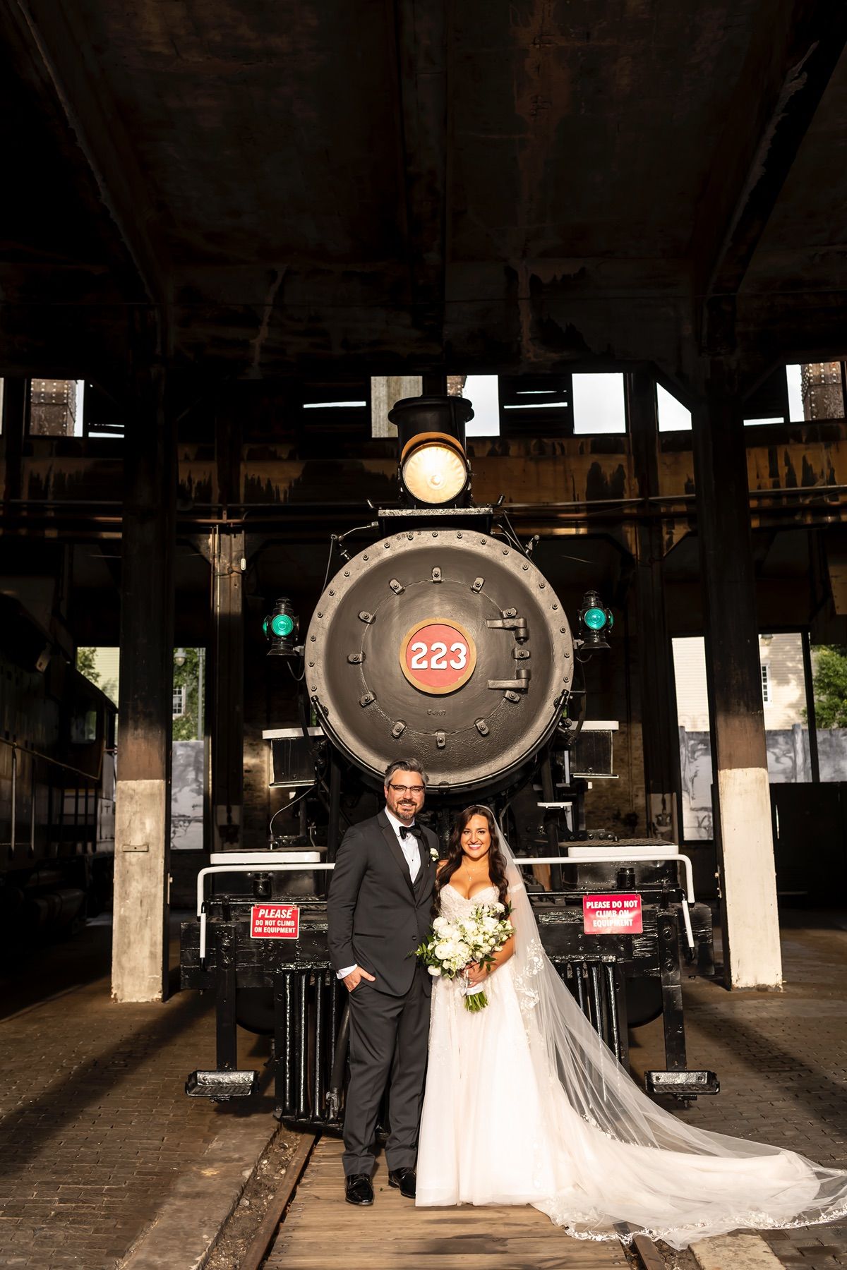 A wedding couple stands in front of a vintage steam locomotive number 223 in a rustic train depot.