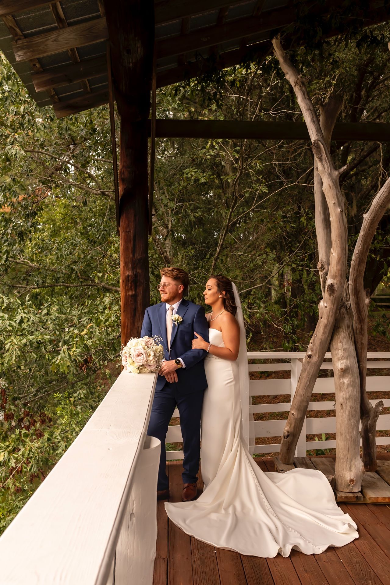 A couple in wedding attire sits together on a wooden bench in an outdoor pavilion surrounded by trees.