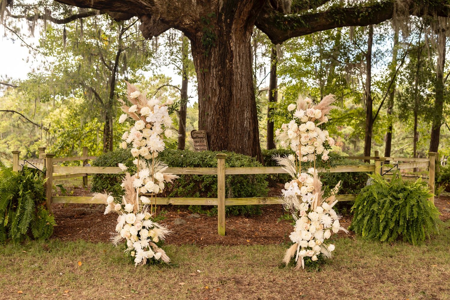 A rustic wooden fence with floral wedding arch under a large oak tree framed by lush greenery in a garden.