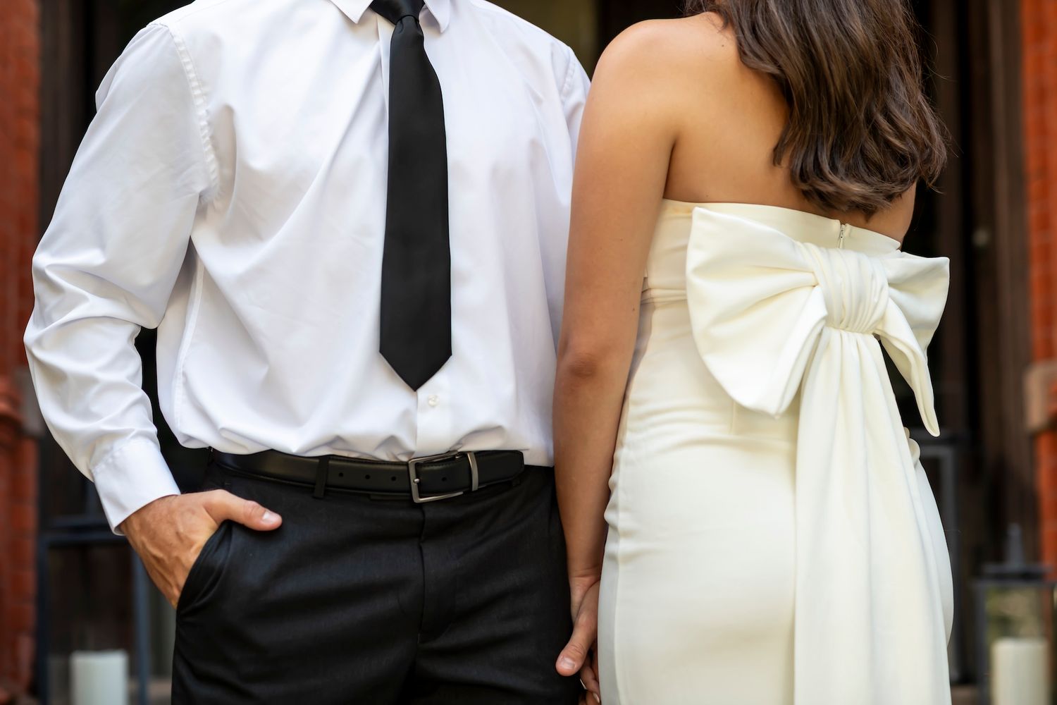 A couple stands together in formal attire with a white dress featuring a large bow and a white shirt with black tie.