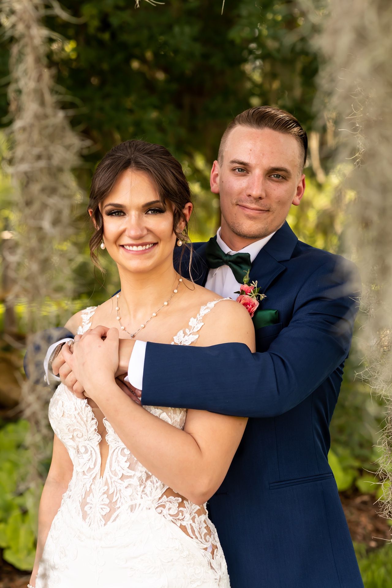 A bride in white lace and groom in navy suit dance together outdoors in a romantic garden setting.