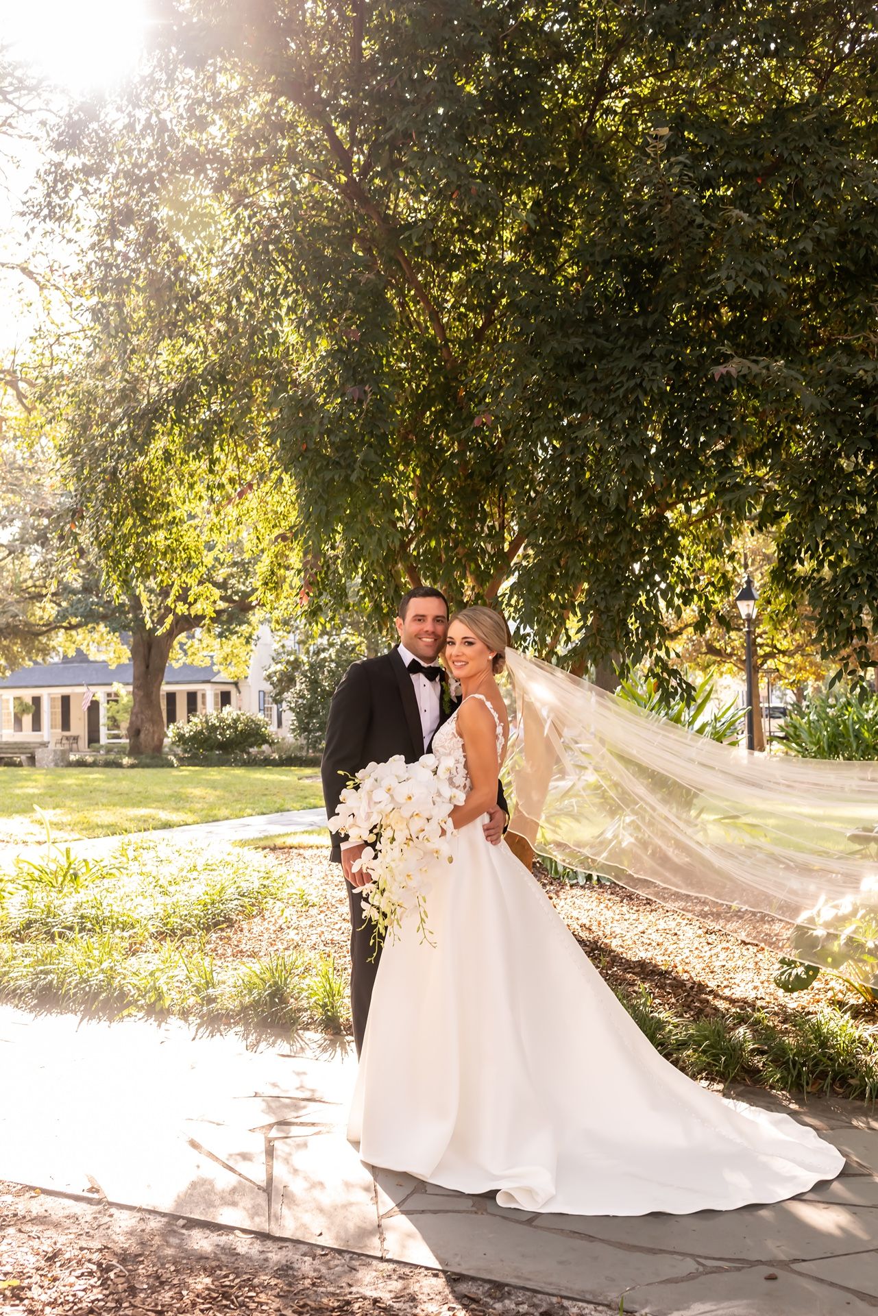 A bride and groom share a romantic moment in a sunny outdoor garden setting as her long veil flows in the breeze.
