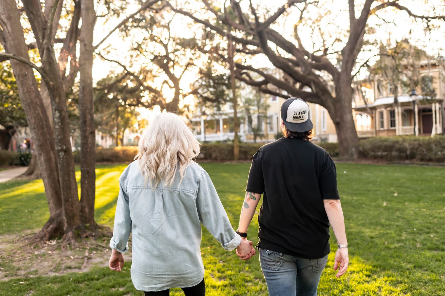 A couple walking hand in hand through a park at sunset with large trees casting shadows on the grass.