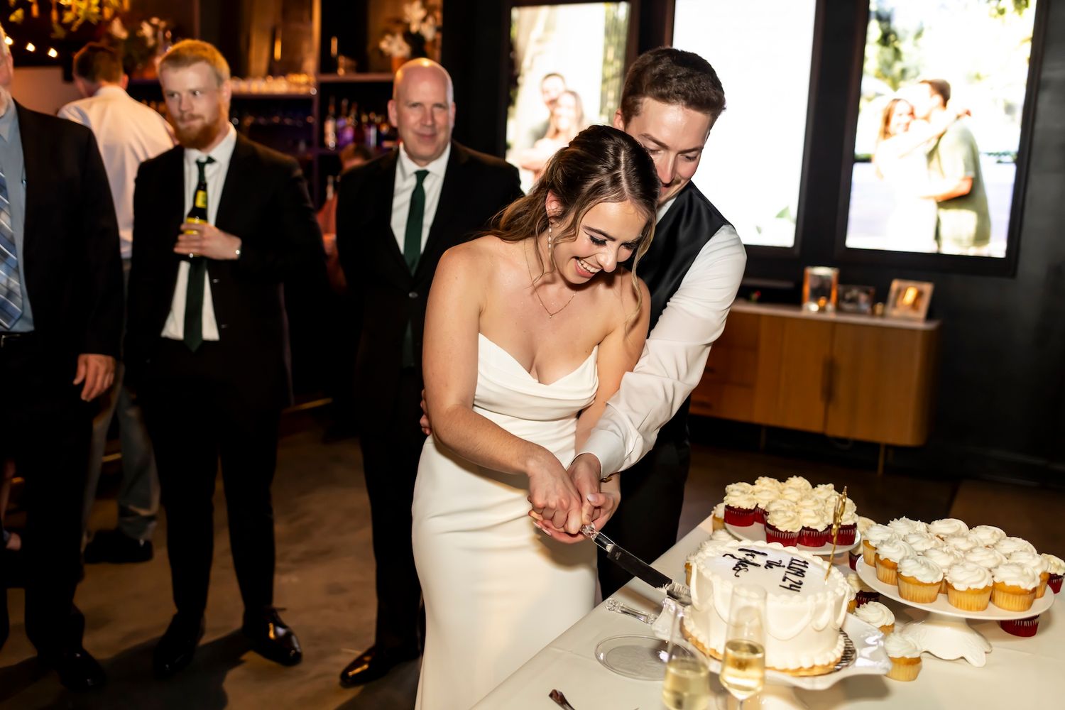 A bride and groom cut their rustic wedding cake together while guests watch at an elegant indoor reception venue.