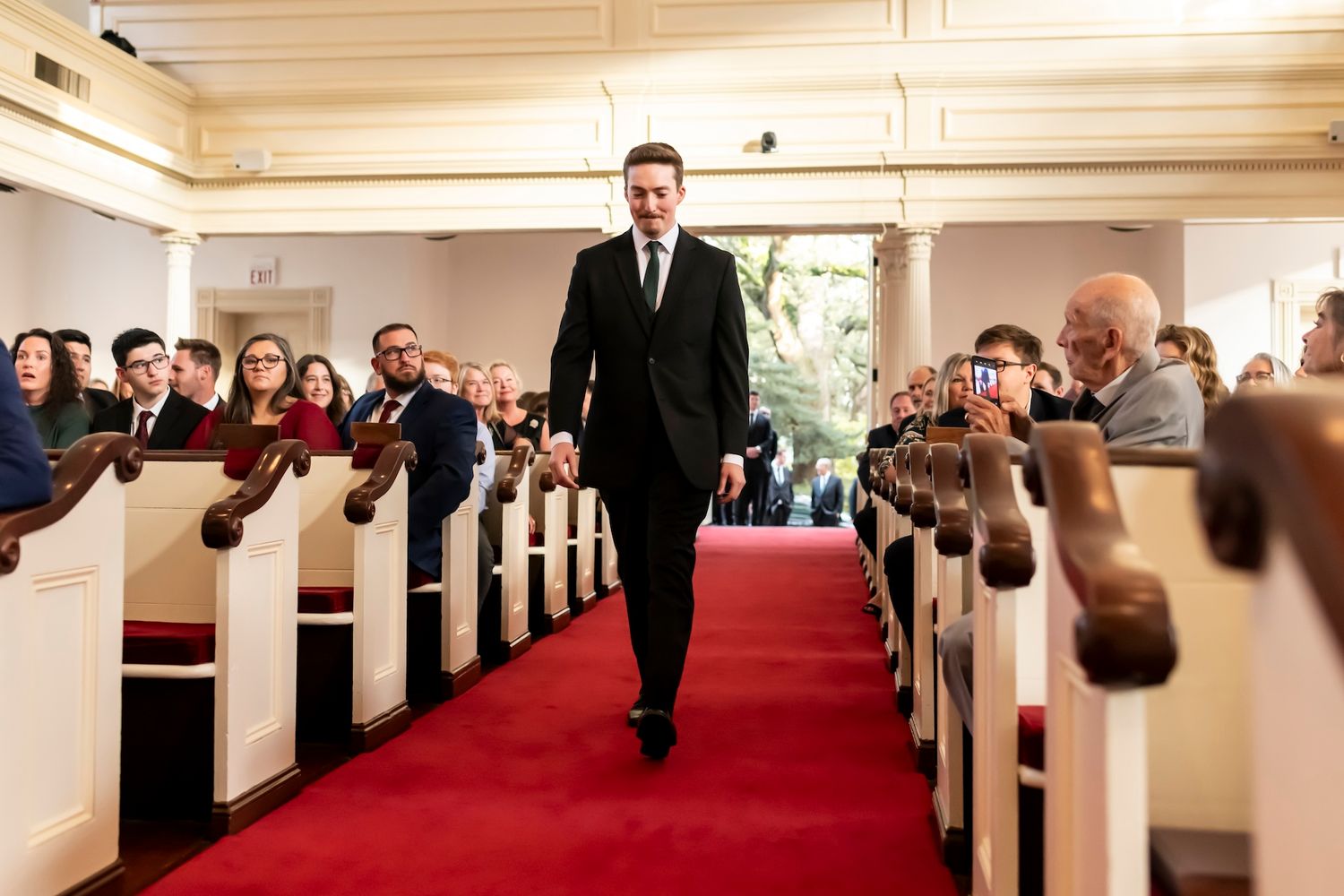 A person in a dark suit walks down the center aisle of a church filled with wedding guests seated in wooden pews.
