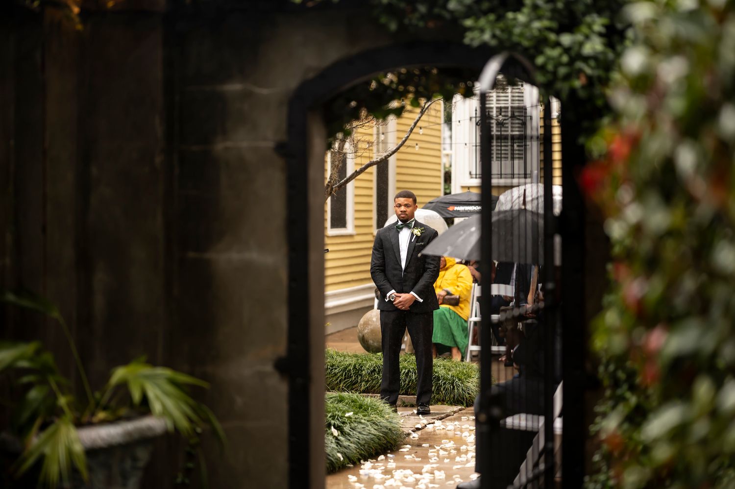 View through ornate iron gate of woman holding bright yellow balloon walking along garden path beside yellow house.