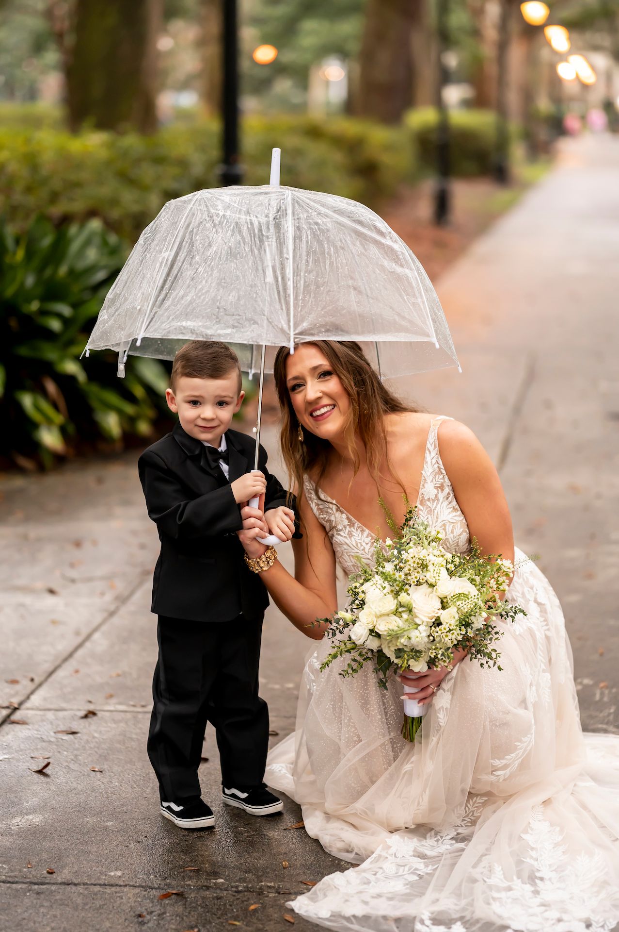 A person in a white lace wedding dress holds a clear umbrella and white bouquet next to a small child in formal attire.