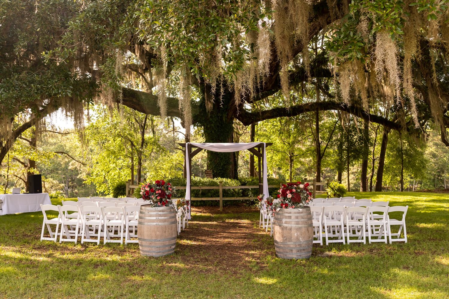 Outdoor wedding ceremony setup with white chairs and wine barrels under Spanish moss draped trees.