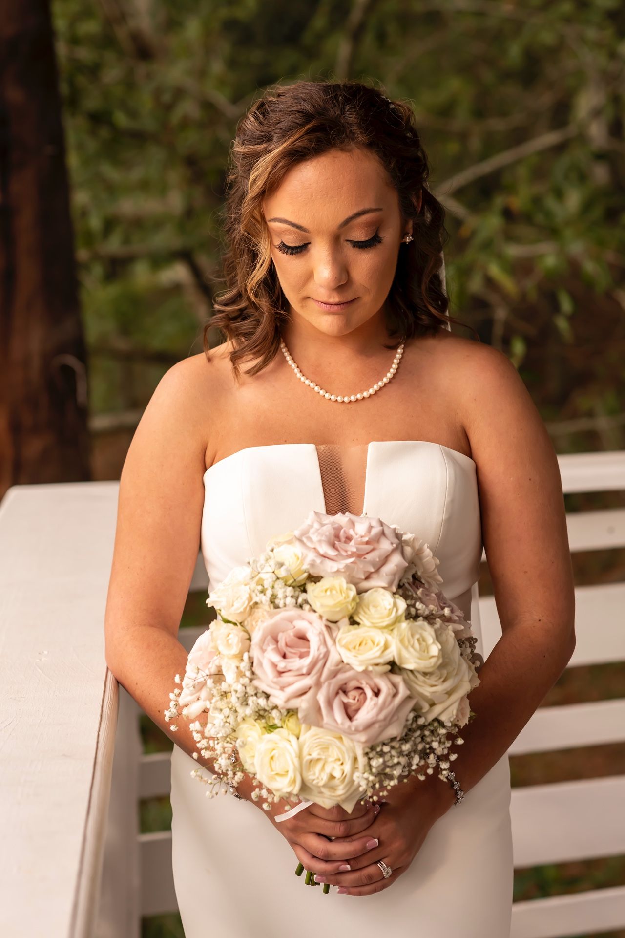 A bride in a white strapless wedding dress holds a beautiful bouquet of pink and cream roses against a white fence.