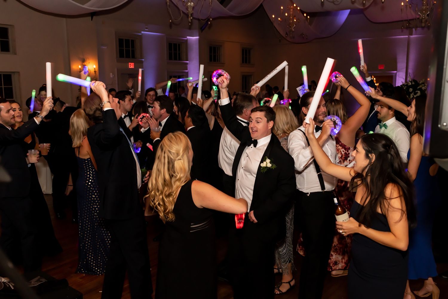 Wedding guests dance while holding up colorful LED light sticks in a dimly lit reception ballroom.