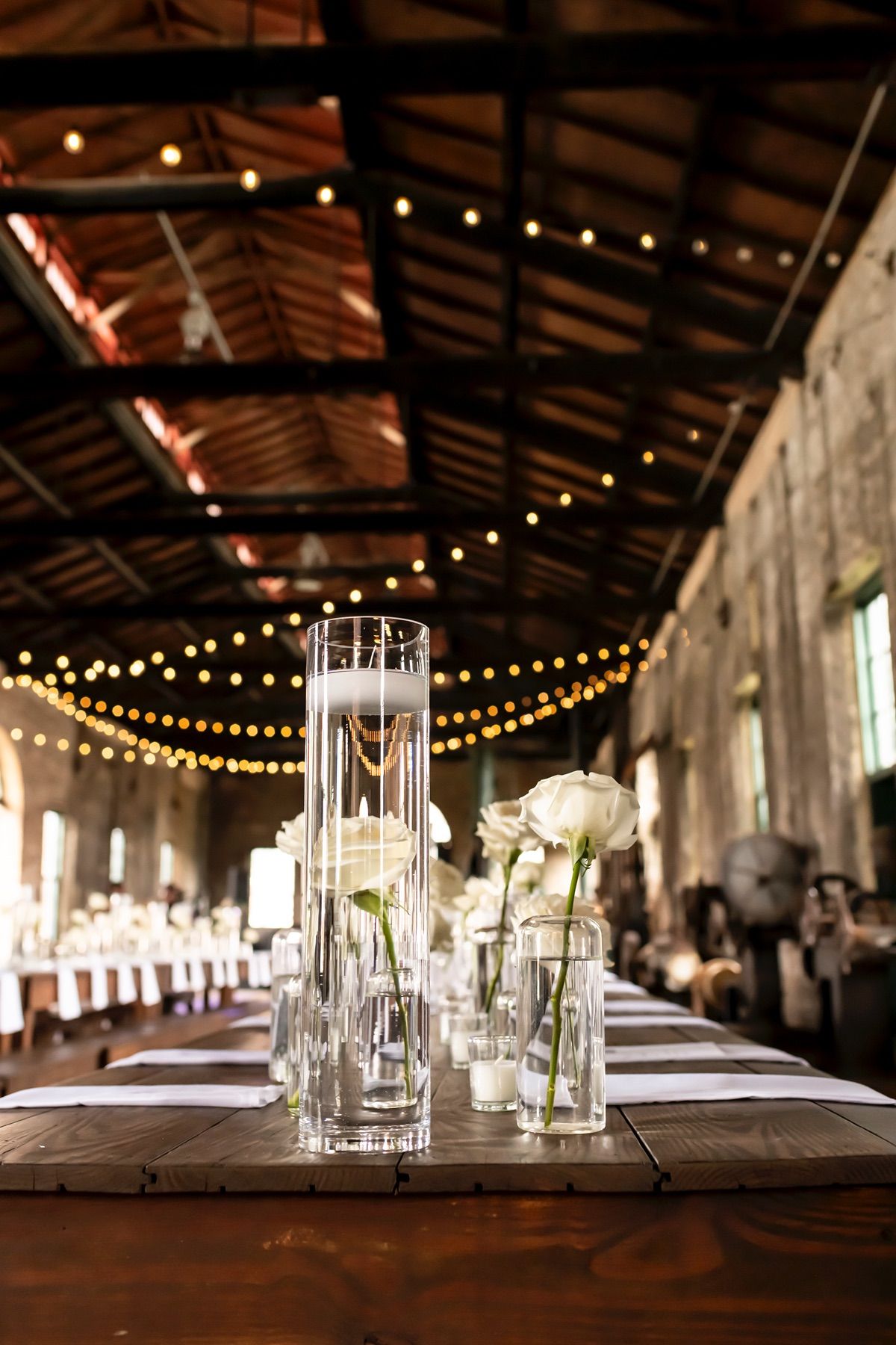 Elegant wedding table decor with glass vases and string lights in a rustic barn venue with exposed beams.