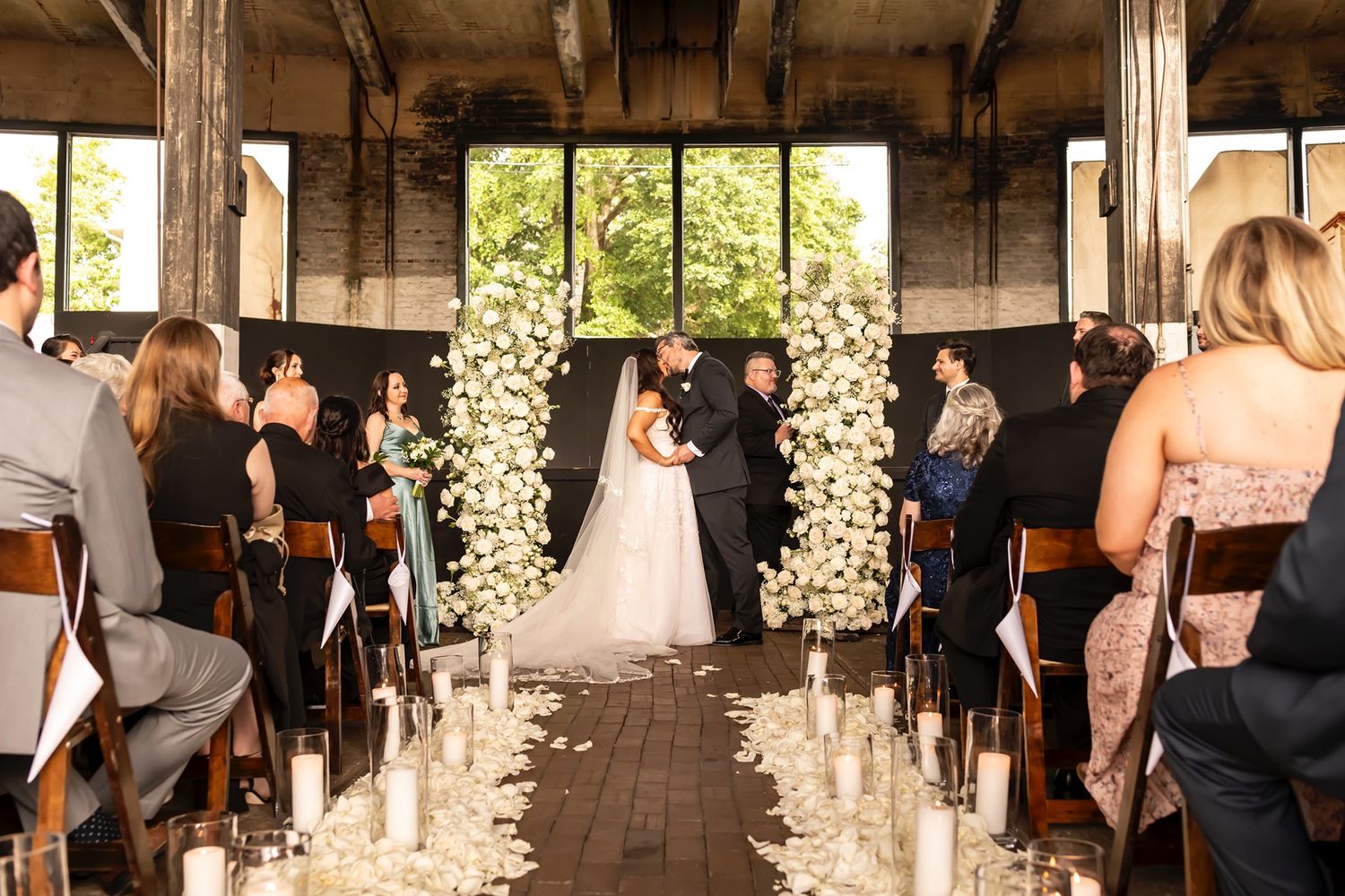 Wedding ceremony takes place in rustic outdoor venue with white floral arch and wooden beams overhead.