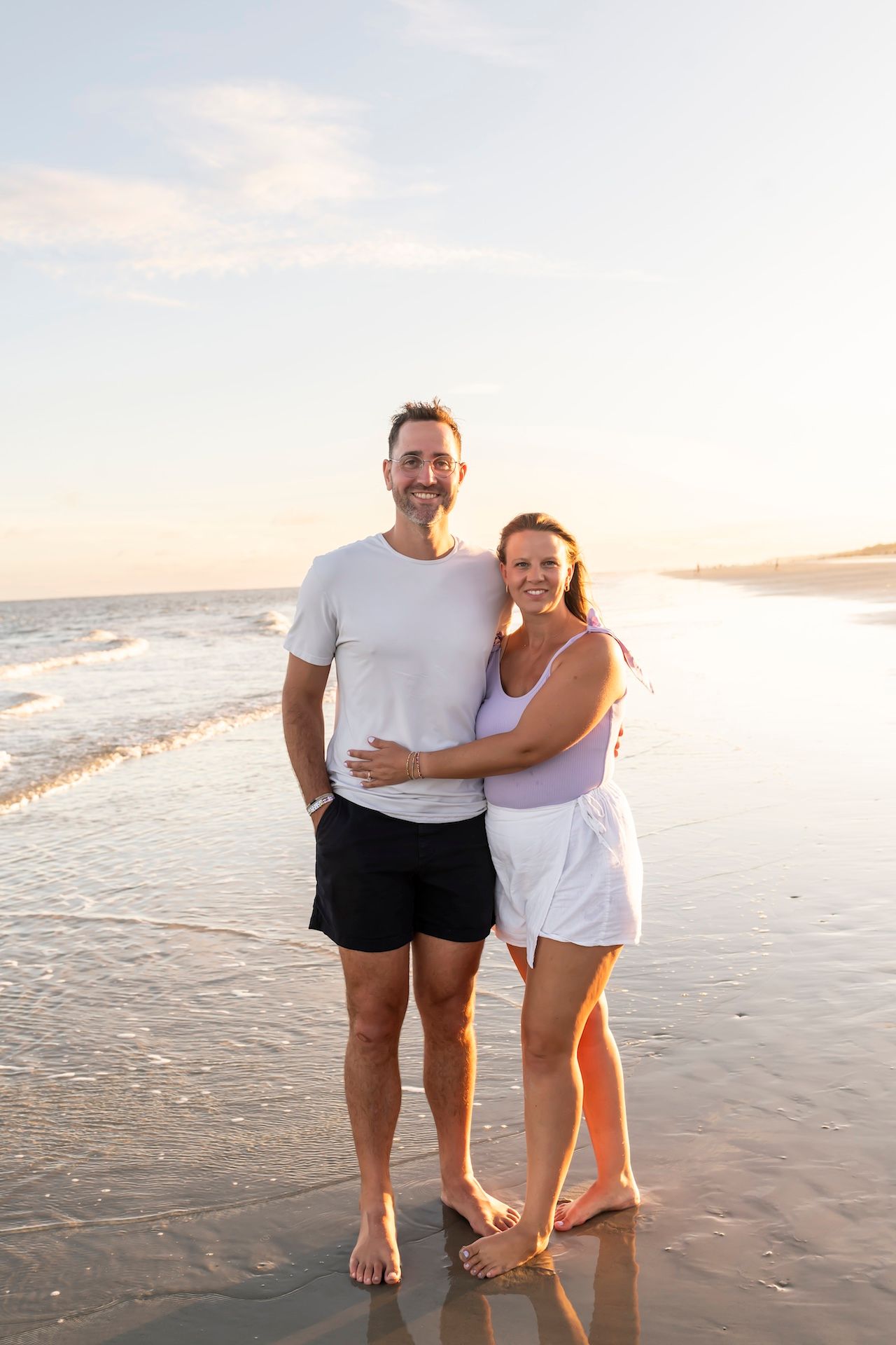 A couple stands together on the beach at sunset with waves gently rolling in behind them.