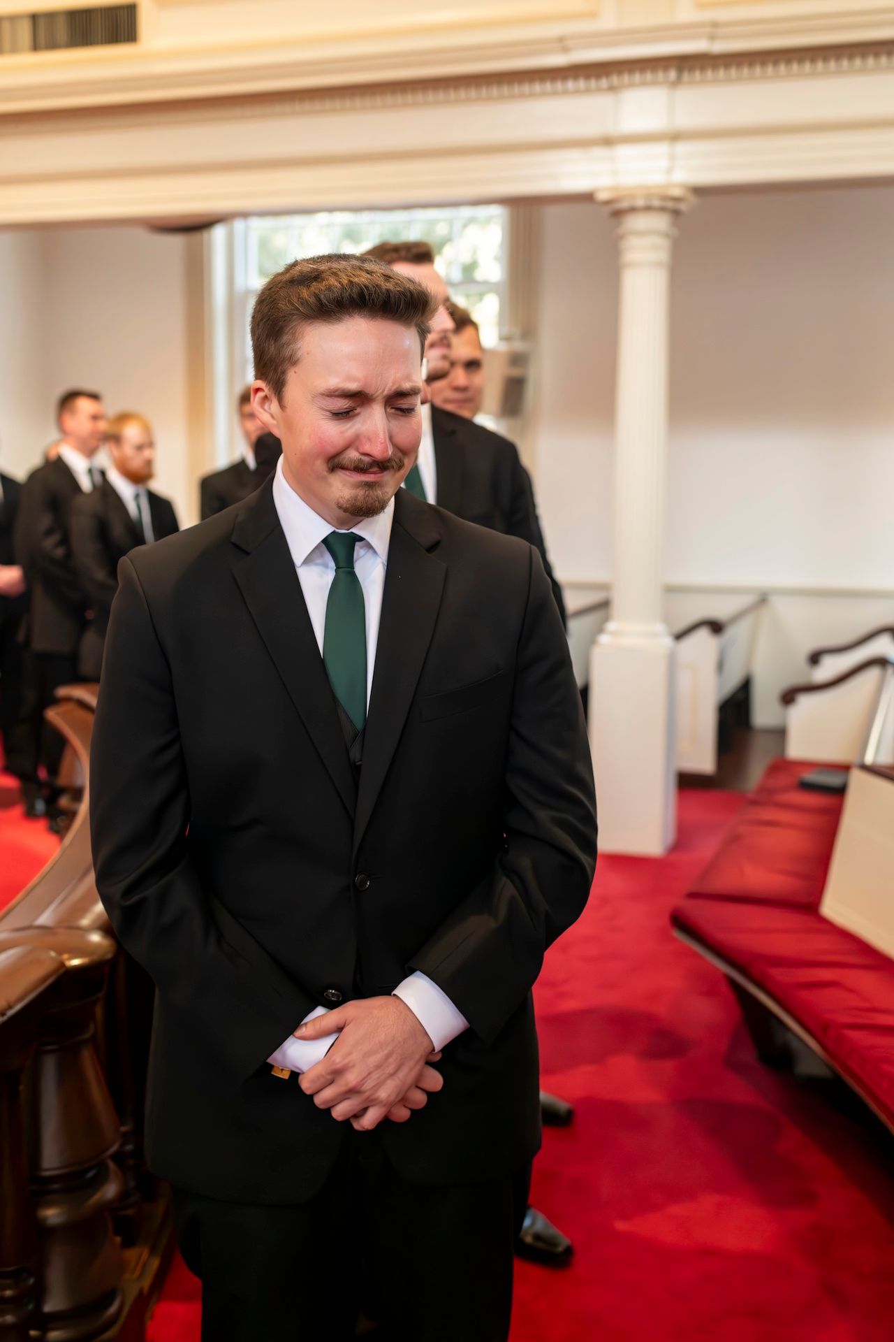 A man in a formal black suit with a green tie stands in an elegant hall with red carpeting and white columns.