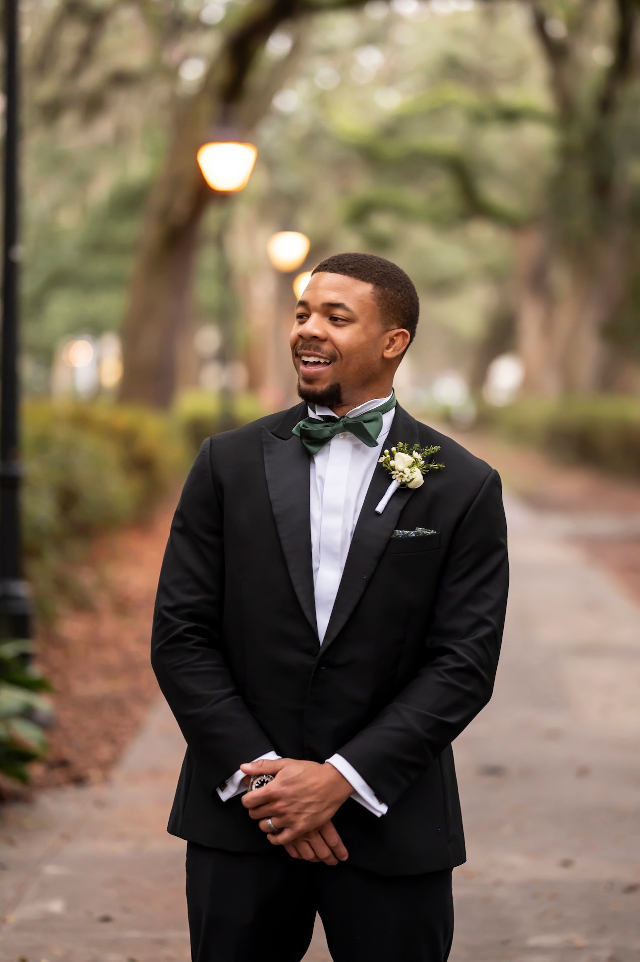A groom in a classic black tuxedo and bow tie smiles joyfully on a tree-lined path with glowing lights in the background.