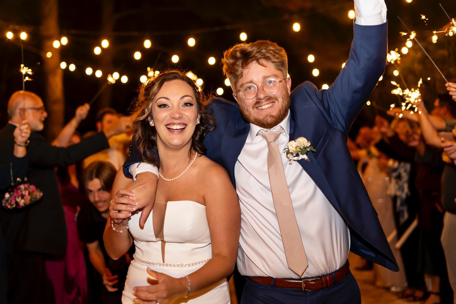 Wedding guests dance and celebrate at a dimly lit evening reception with string lights glowing overhead.