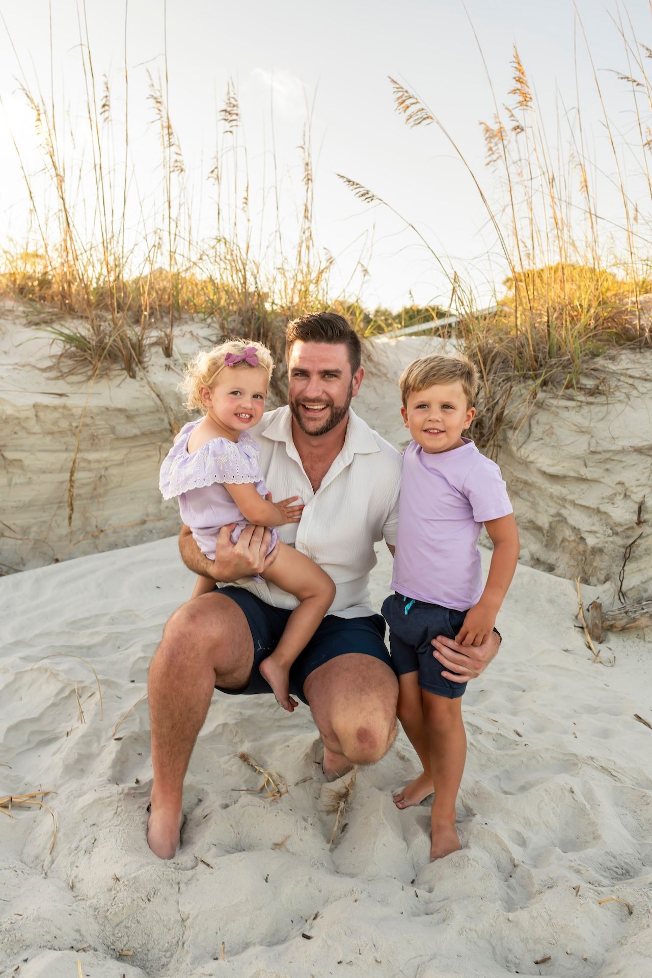 A tender beach portrait of a family sitting together on sandy dunes with sea oats swaying in the golden sunlight.