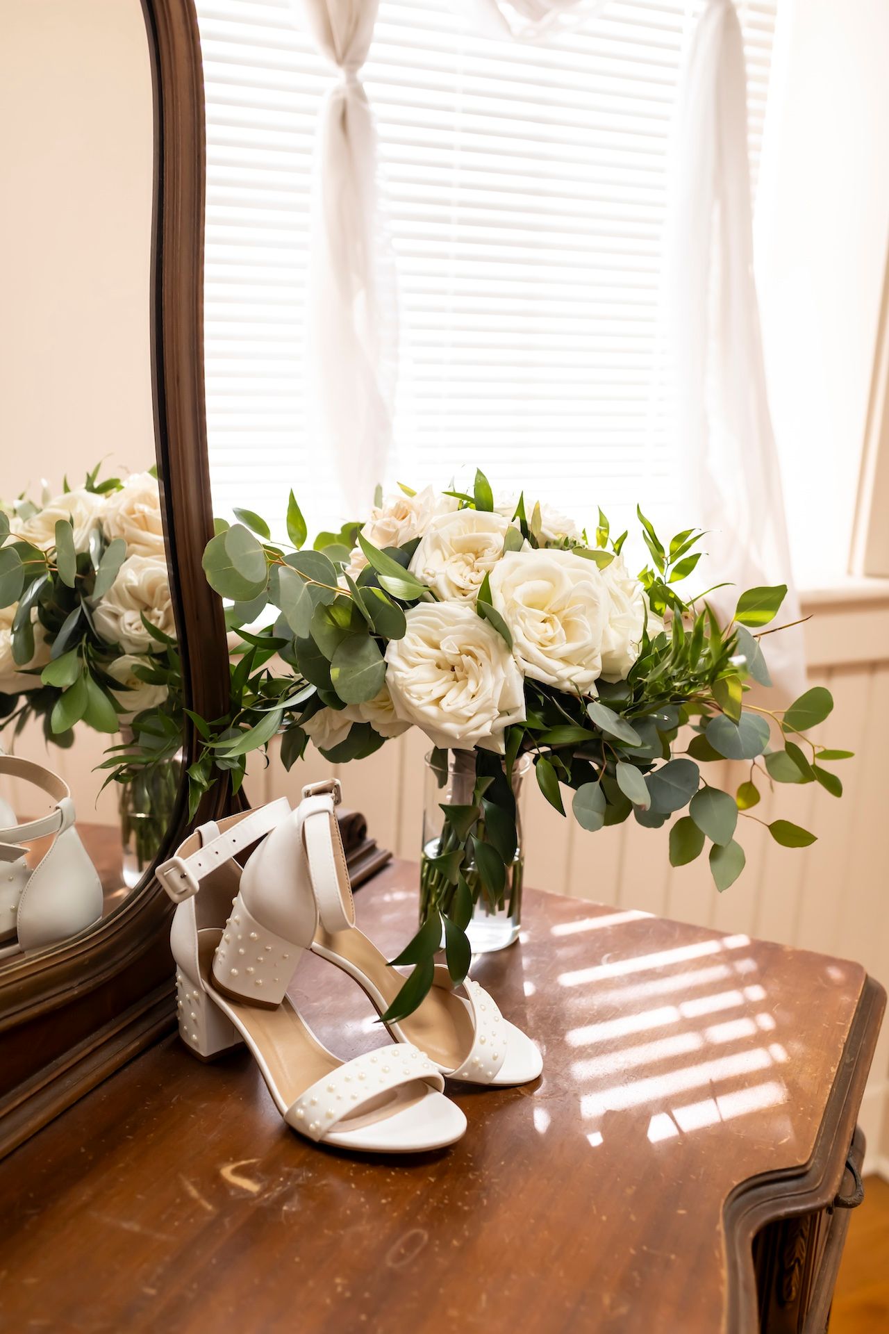 Elegant white wedding roses and greenery arrangement displayed with silver heels on a vintage wooden dresser.