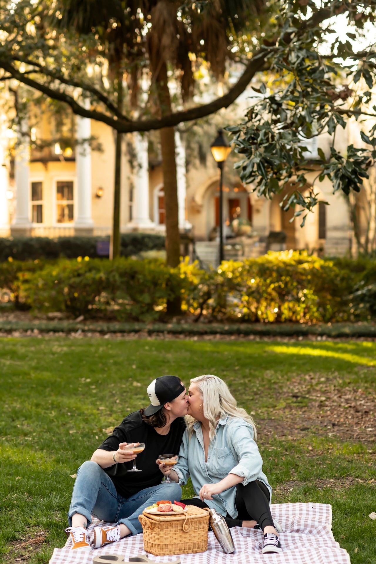 Couple enjoying romantic sunset picnic on blanket with wine and picnic basket in park setting.