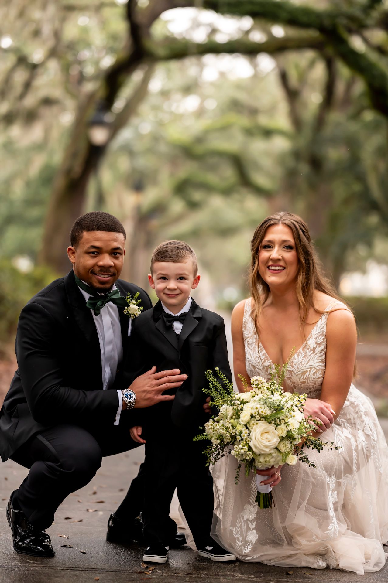 A young happy couple in wedding attire poses with an excited child on a tree-lined path.