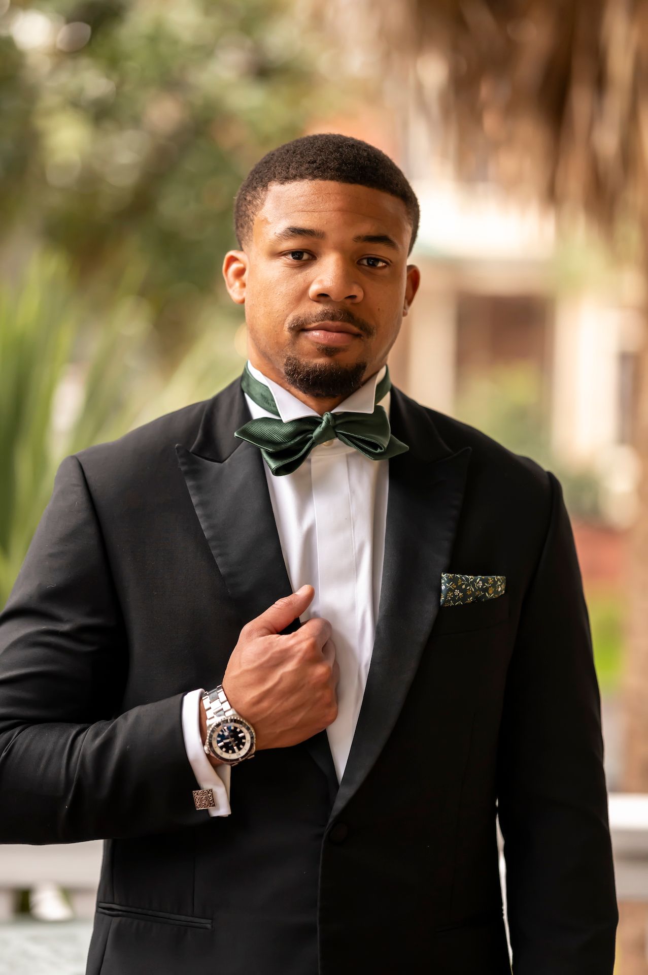 Formal portrait of someone wearing a black tuxedo with green bowtie and pocket square while adjusting their watch.