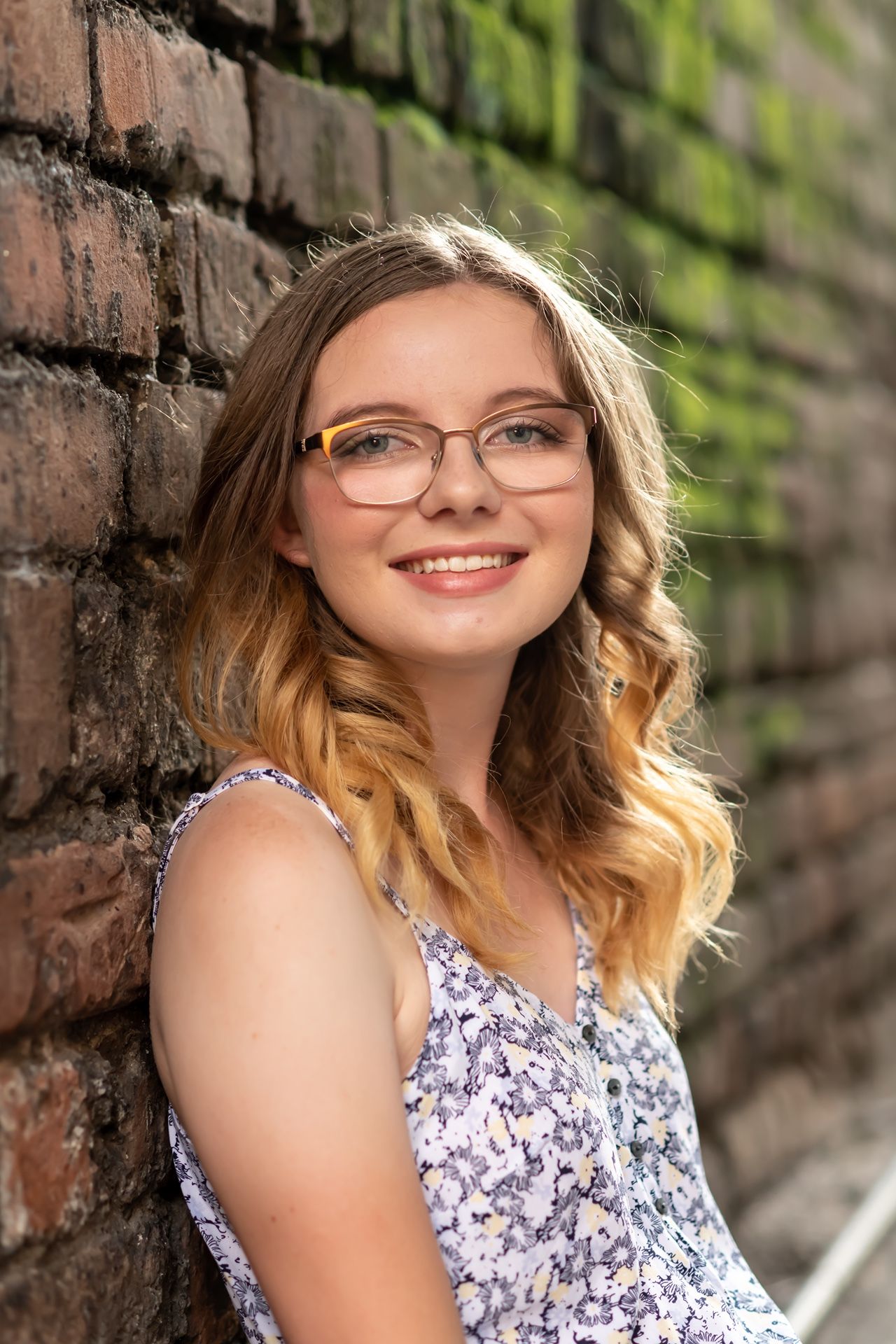 Portrait photographs in a summery outdoor setting with brick wall backdrop featuring a floral dress and eyewear.
