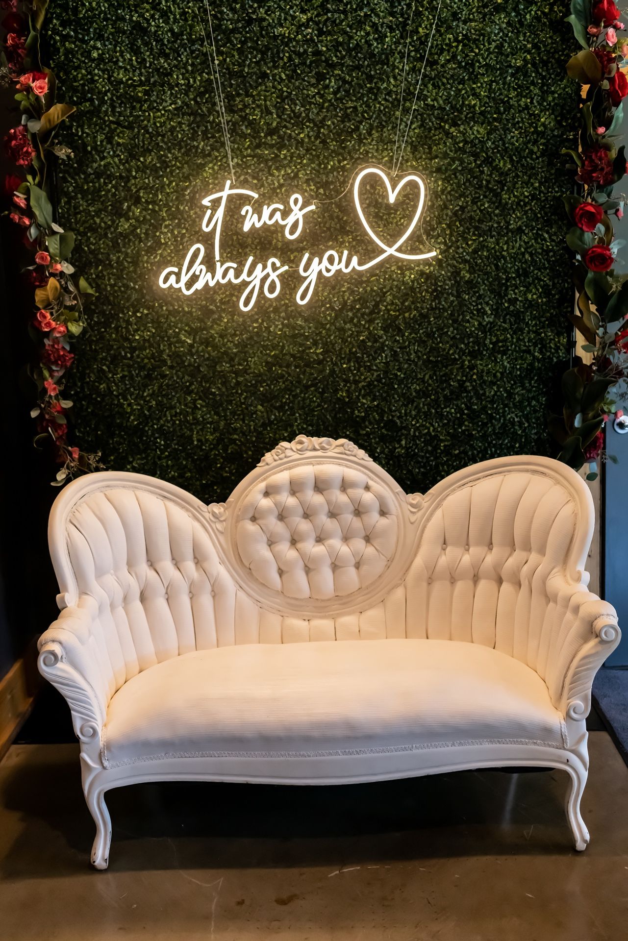 Elegant vintage white tufted sofa with neon 'I was always you' heart sign against a green hedge wall backdrop.