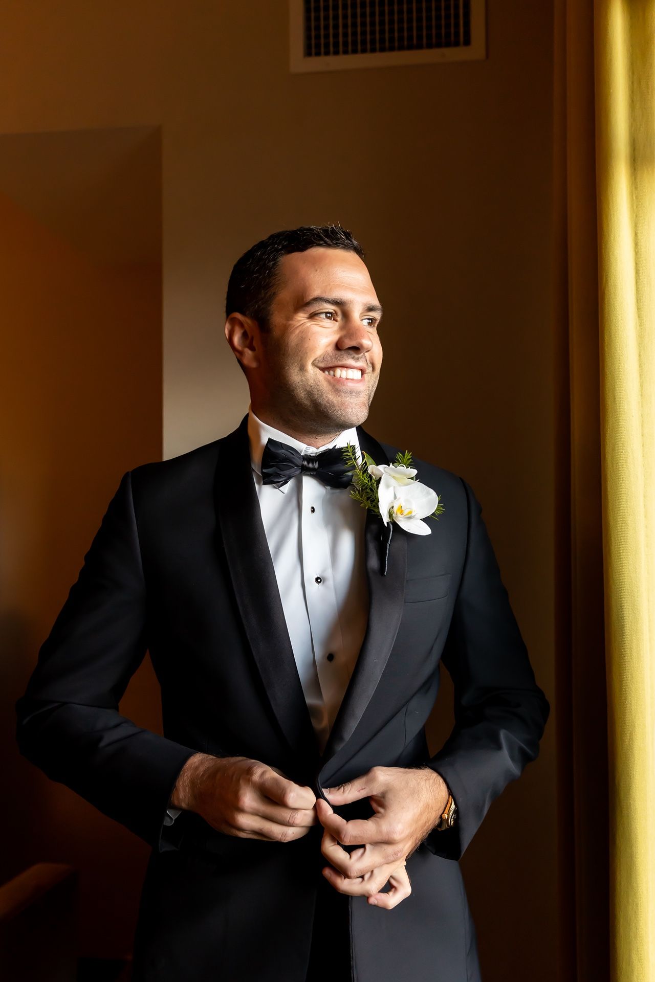 A groom in a classic black tuxedo and white boutonniere adjusts his bowtie in warm ambient lighting.