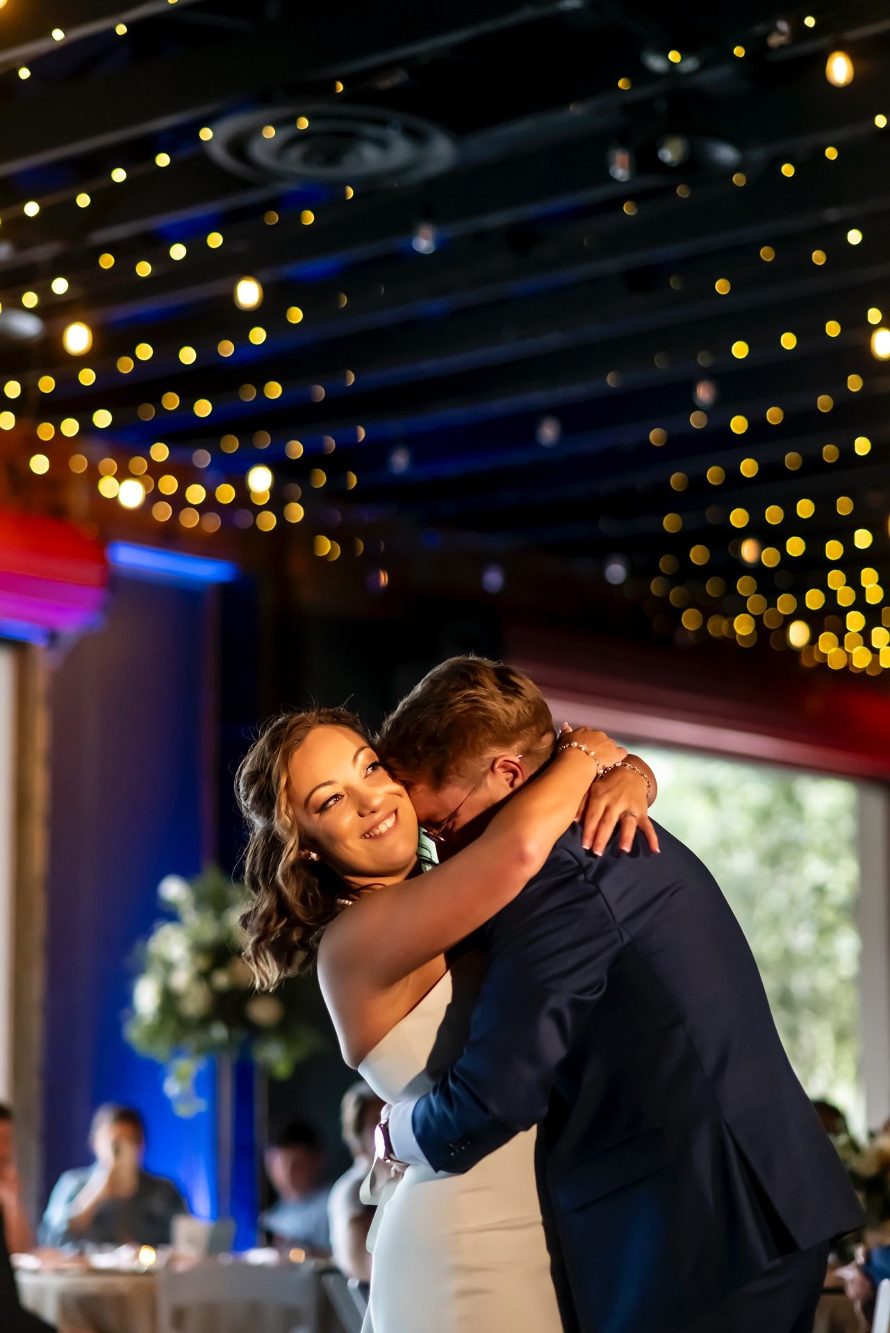 Couple embraces during romantic first dance at wedding reception under twinkling string lights.