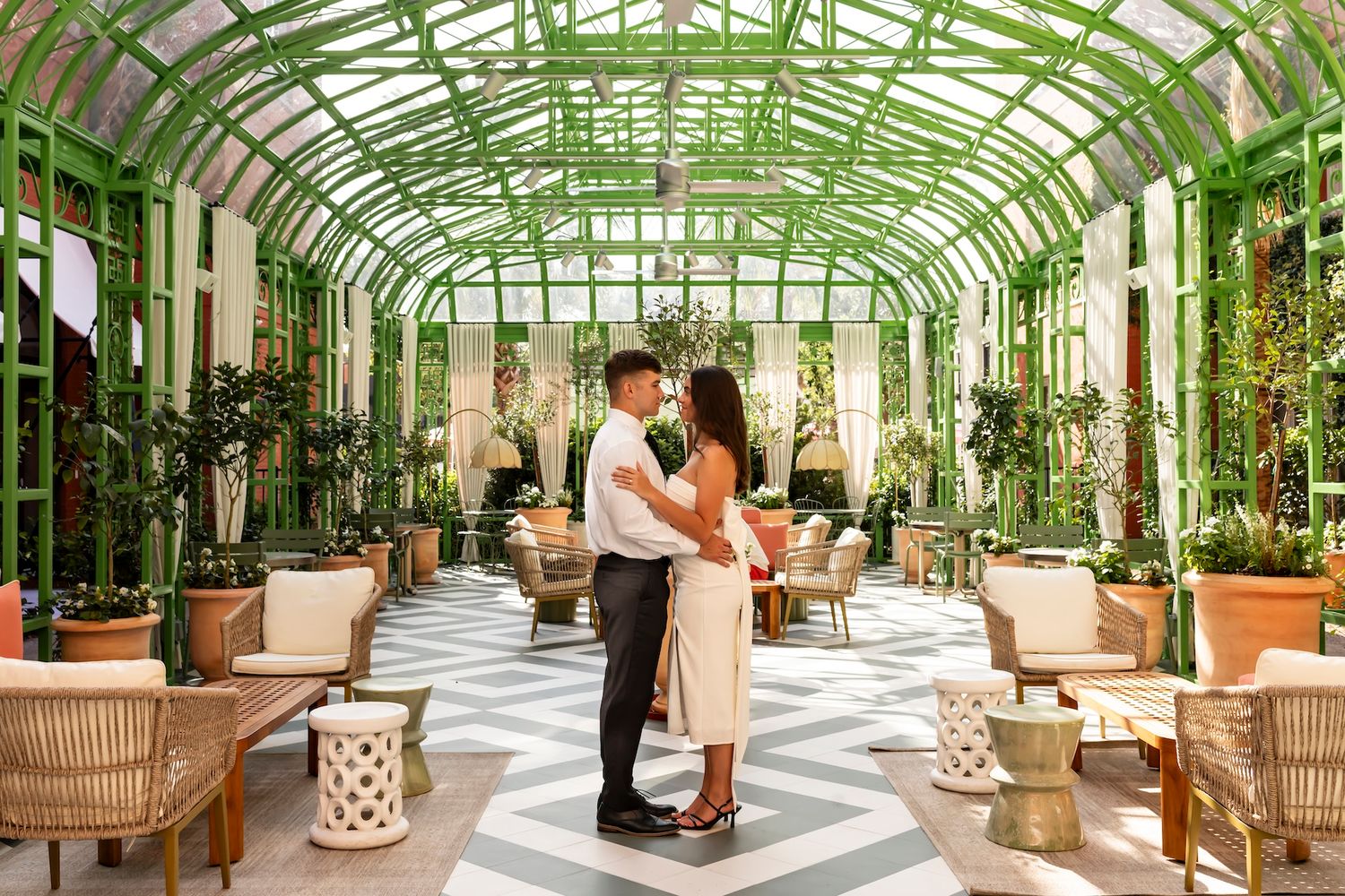 A couple embraces in a stunning greenhouse conservatory with vaulted glass ceilings and elegant furniture.