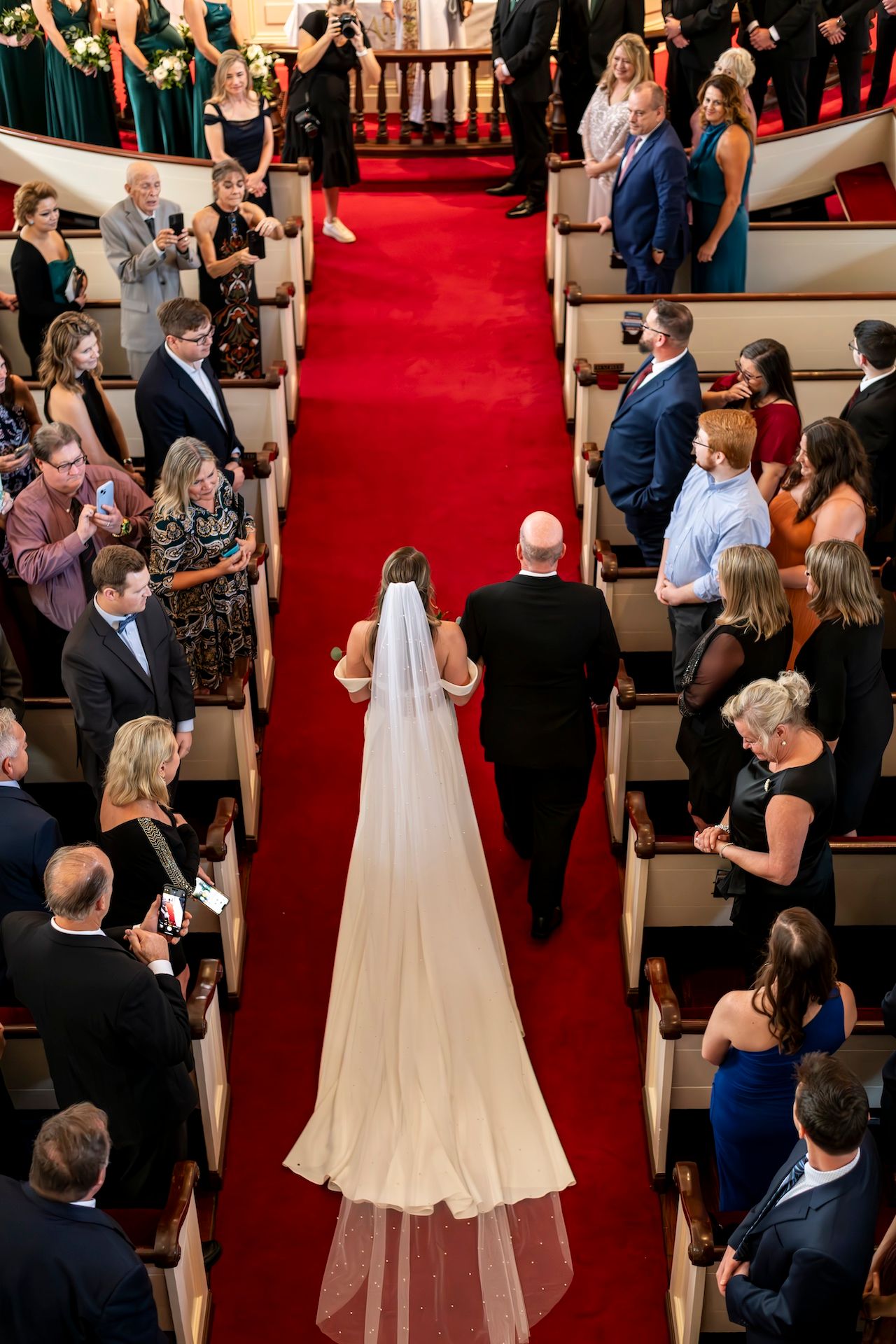 Overhead view of wedding ceremony in church with long flowing veil trailing down red carpeted aisle surrounded by guests.
