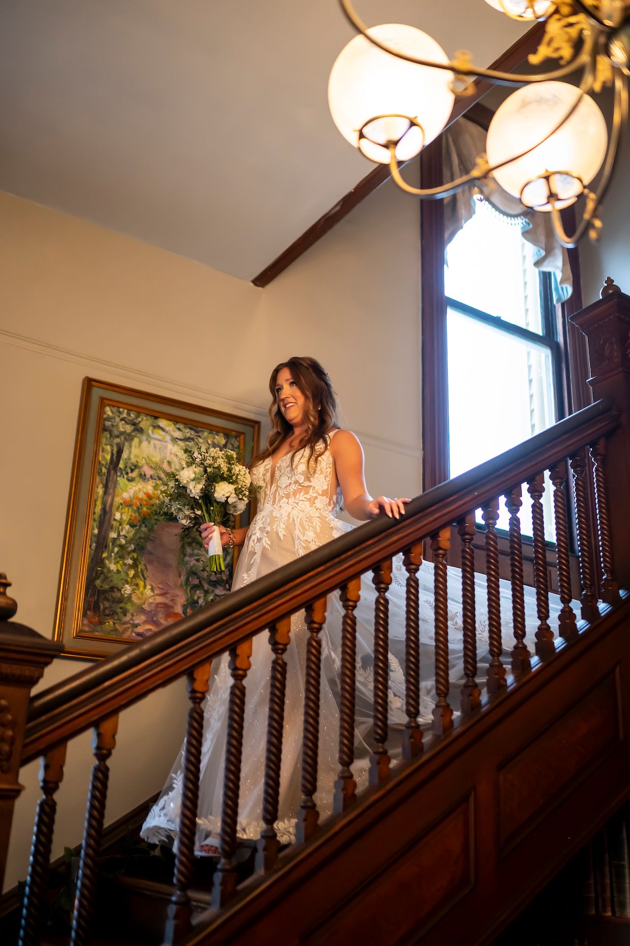 Person in a lace gown descends an elegant wooden staircase under warm lighting during a special event.