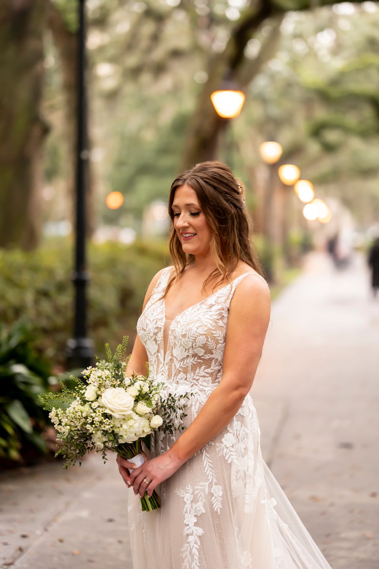 A bride in a white lace gown holds a bouquet of white flowers while standing outdoors on a pathway lit by string lights.
