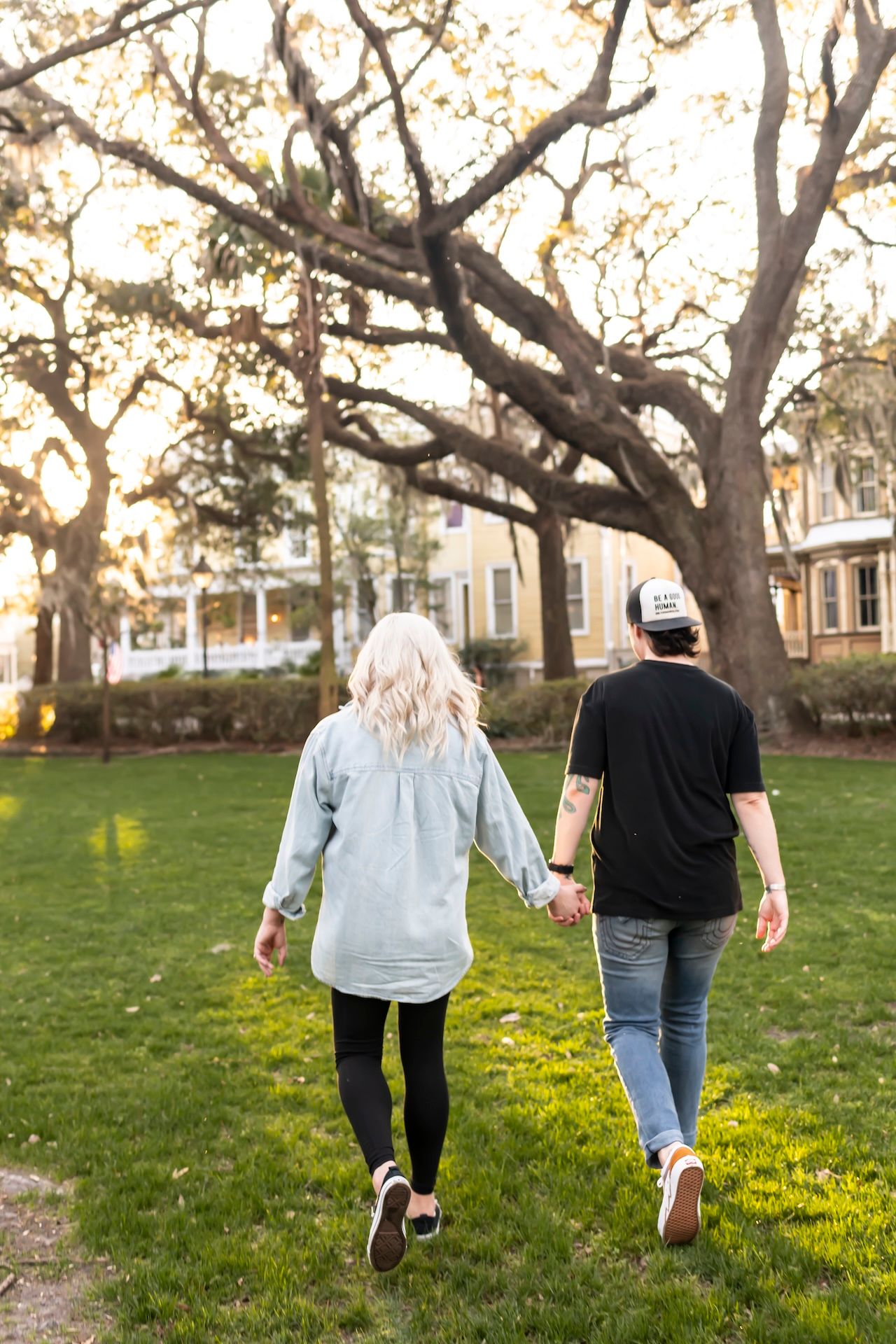 Two people walking on grass holding wine glasses at sunset, taken from behind under autumn trees.
