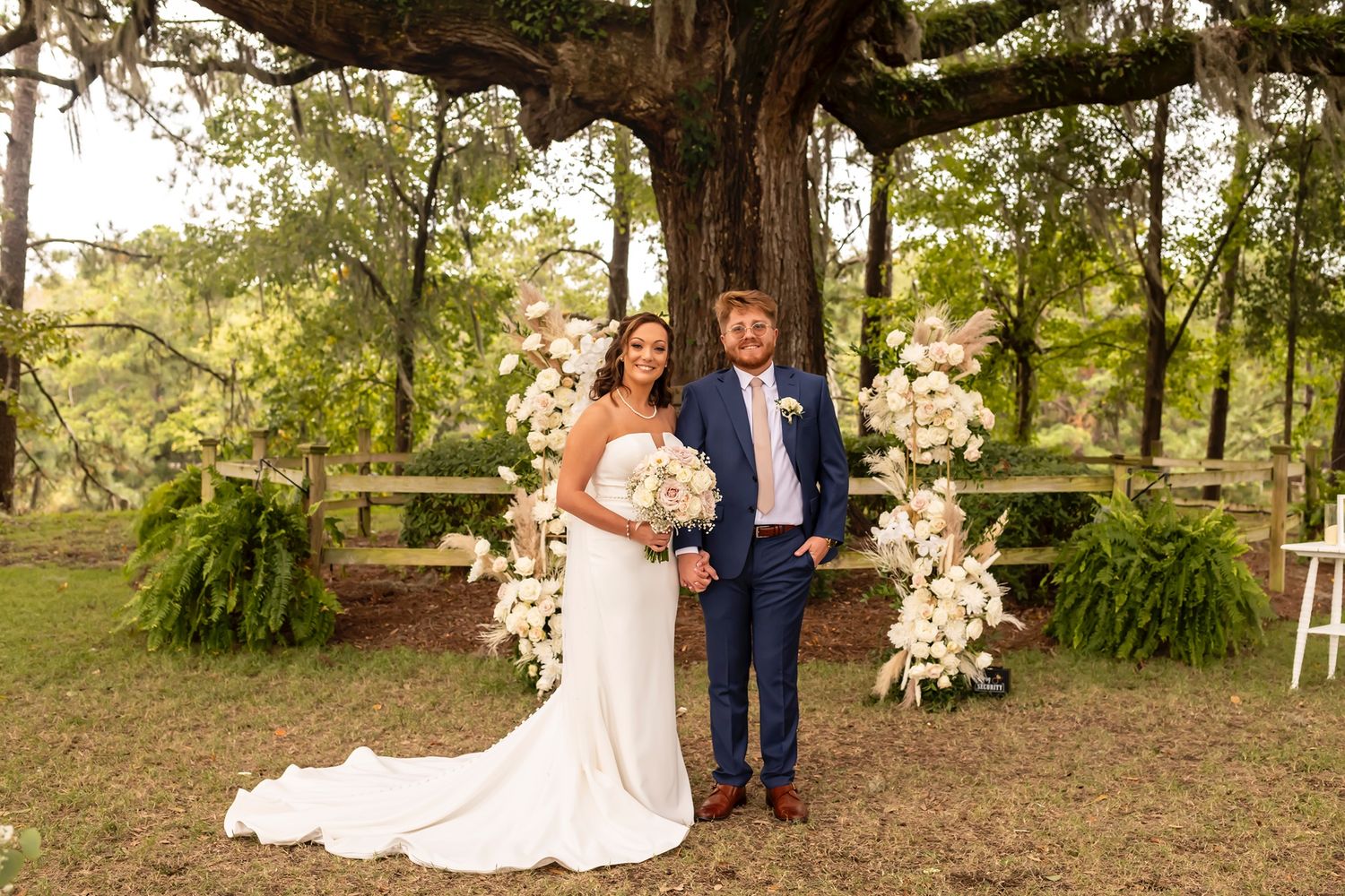 Wedding couple poses under a majestic oak tree decorated with white floral arrangements in a rustic outdoor setting.