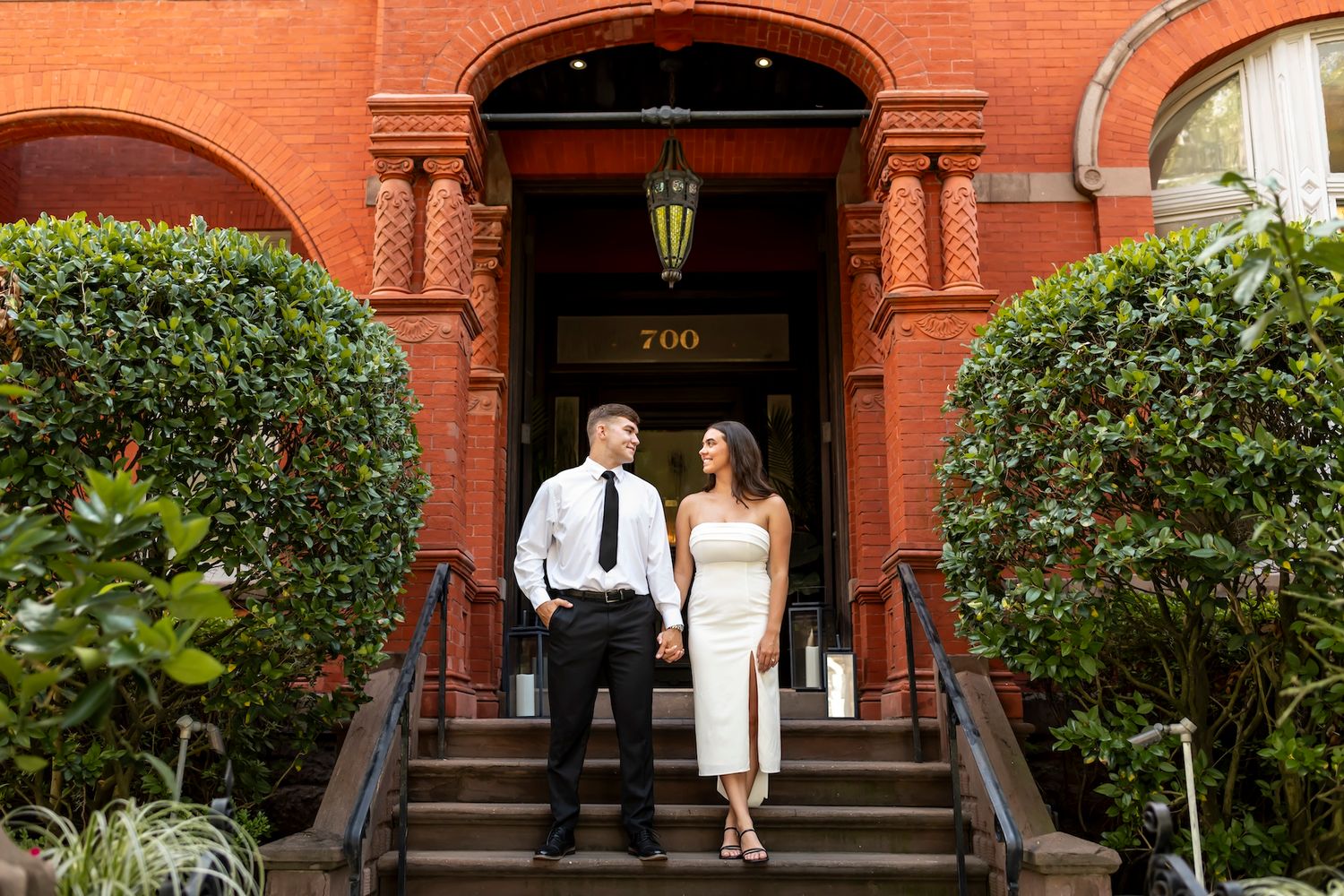 A couple stands on the front steps of a historic red brick building with lush greenery flanking the entrance.