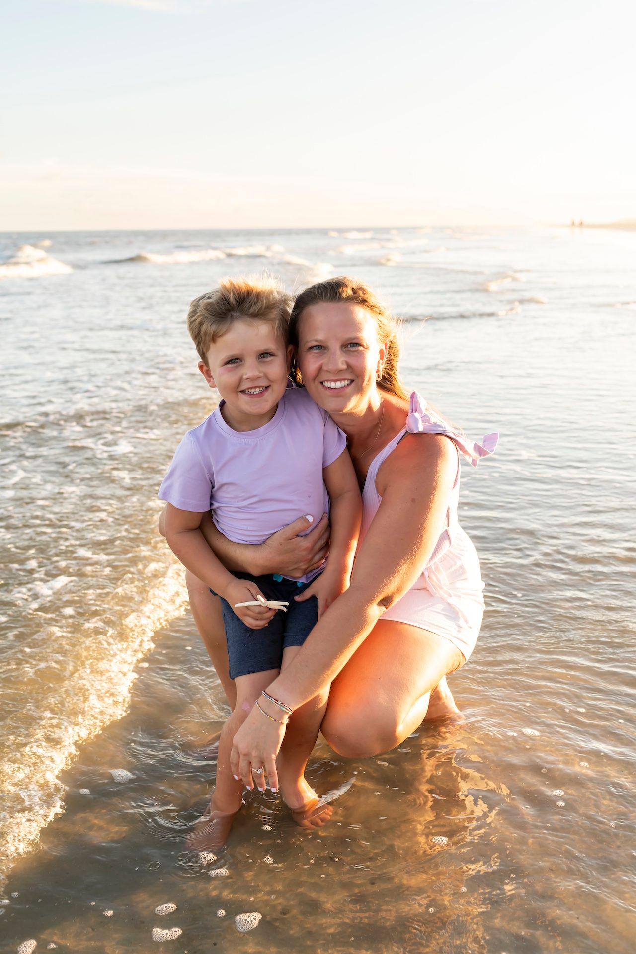 Two people share a joyful embrace while sitting in shallow ocean waves at sunset.