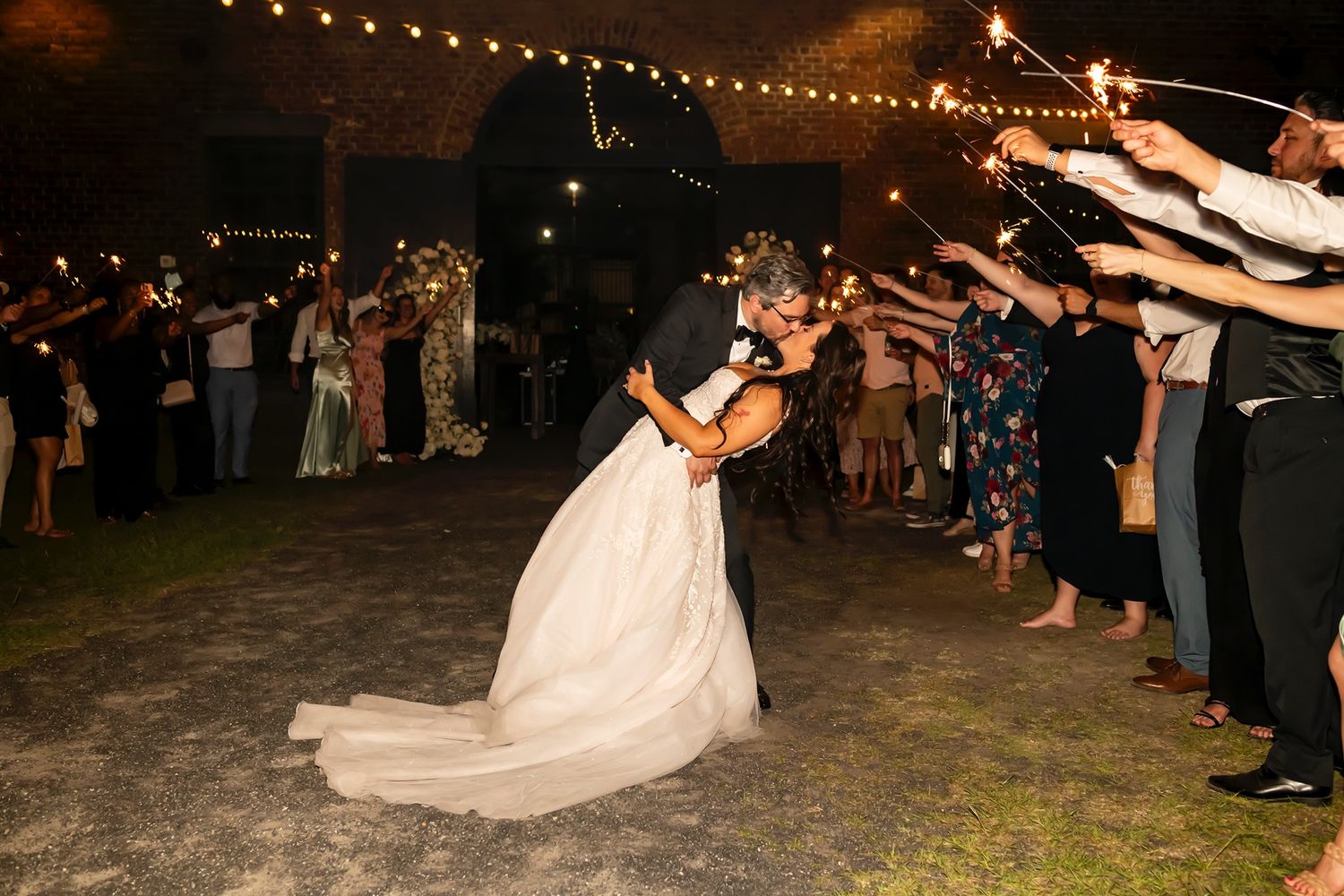 Couple shares romantic dip kiss during sparkler wedding exit while guests hold sparklers forming illuminated archway at night.
