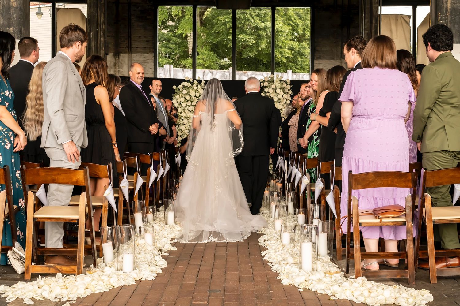 Wedding ceremony processional along brick aisle lined with glowing candles while guests watch from wooden chairs.