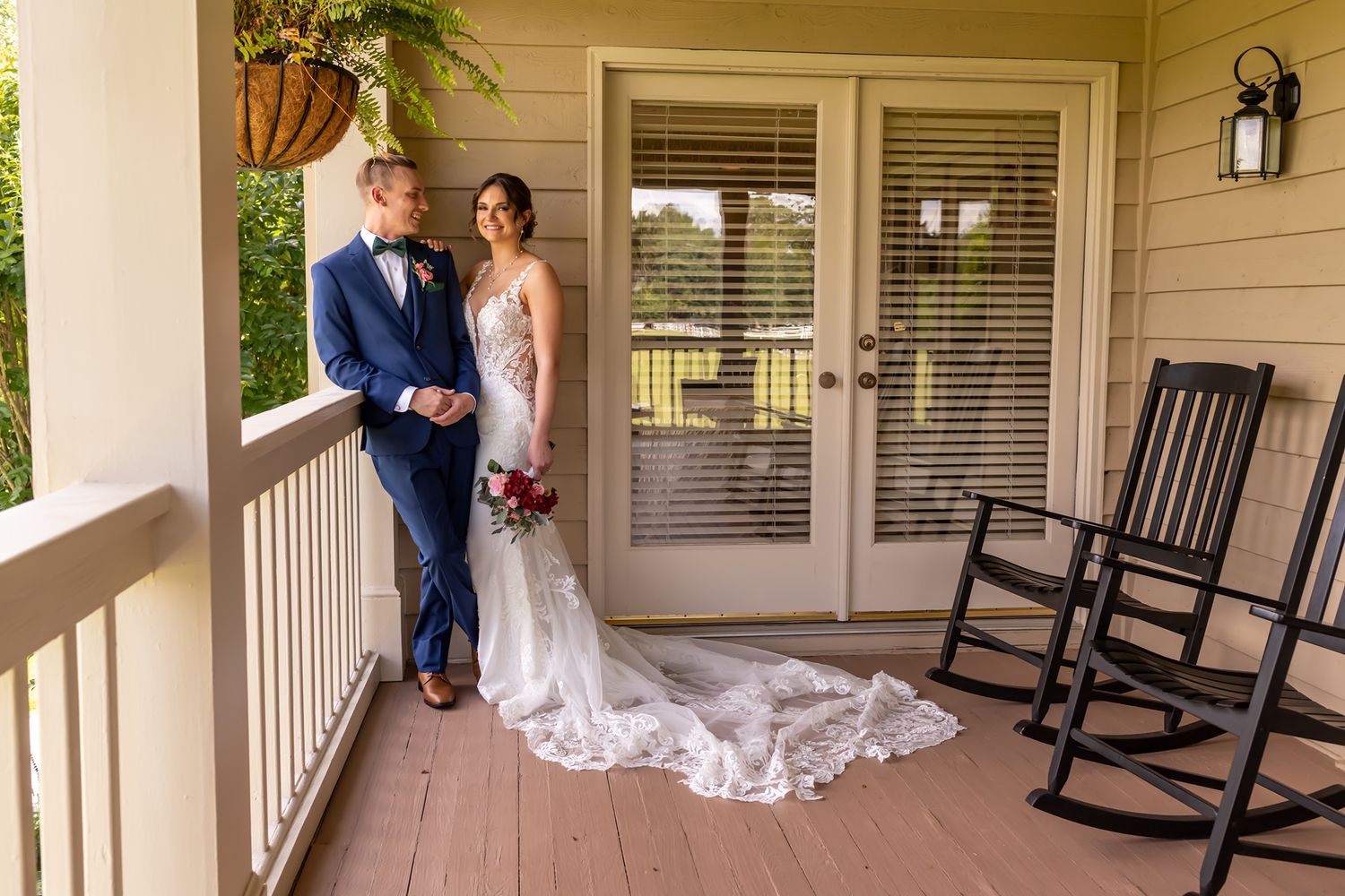 A couple stands on a covered porch with wooden rocking chairs and beautiful white dress with long flowing train.