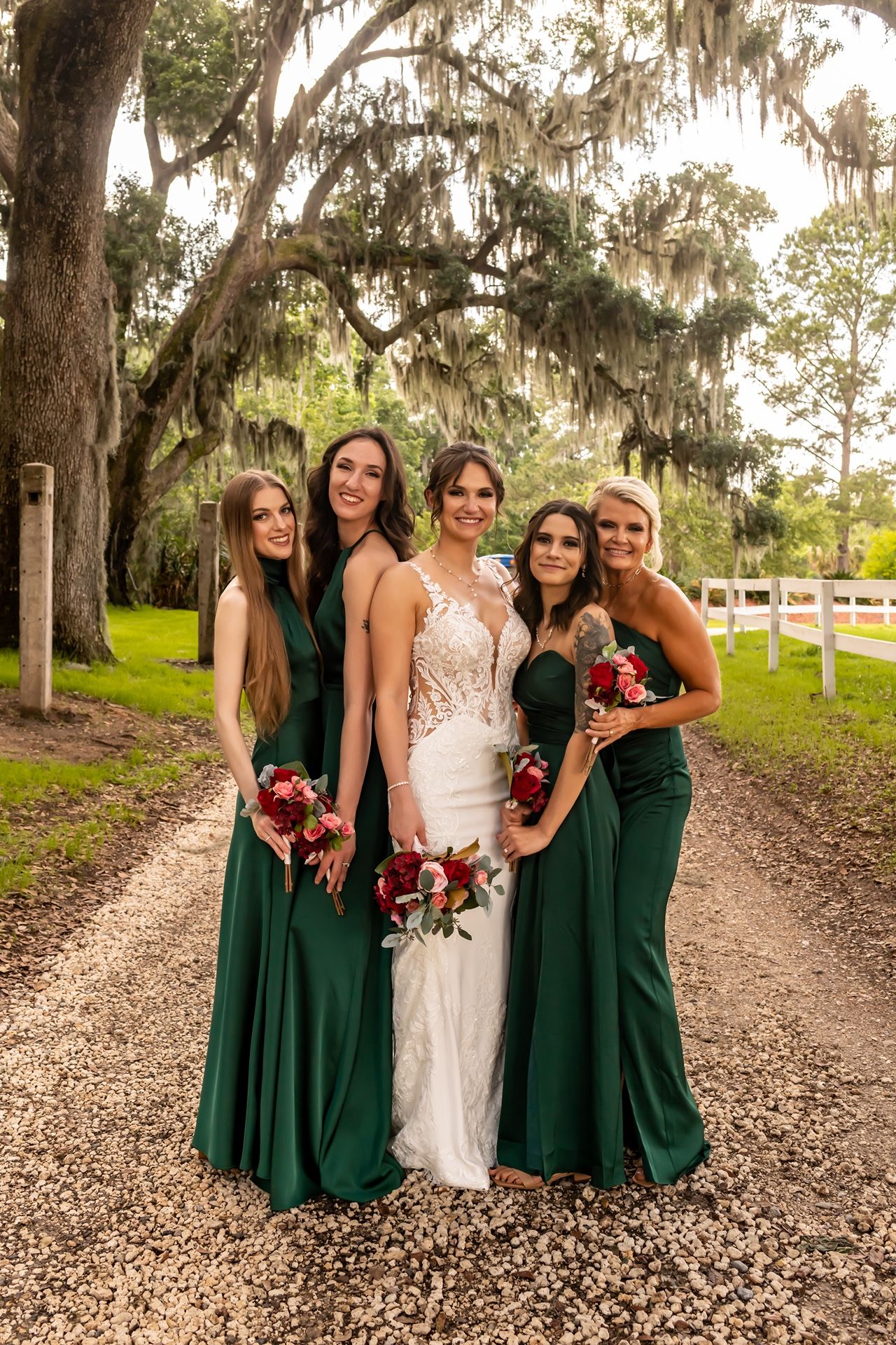 Wedding party wearing elegant emerald green bridesmaid dresses and white lace bridal gown pose on rustic path under moss-draped trees.