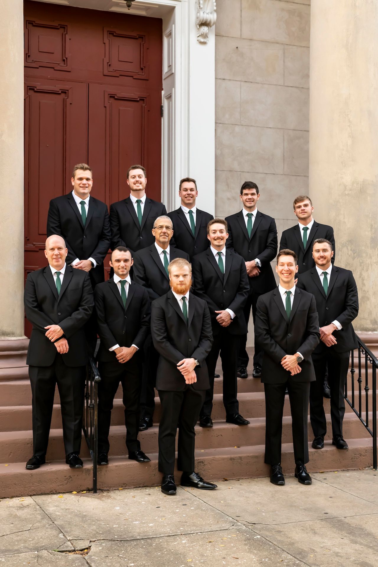 Groomsmen in black suits with green ties pose on stone steps in front of a burgundy door.