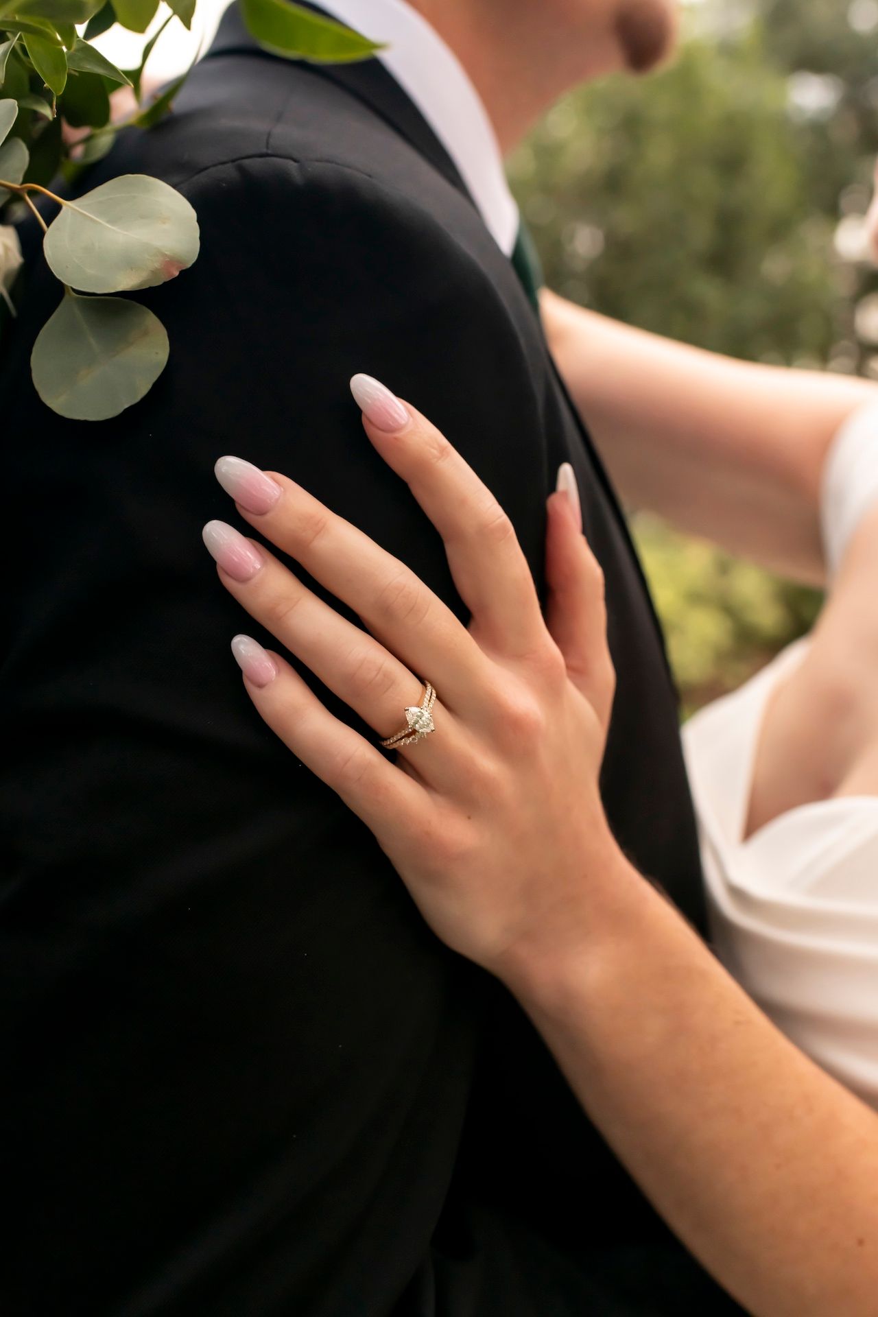 A close-up shot of a bride's hand with engagement ring resting on groom's black suit jacket with eucalyptus leaves.